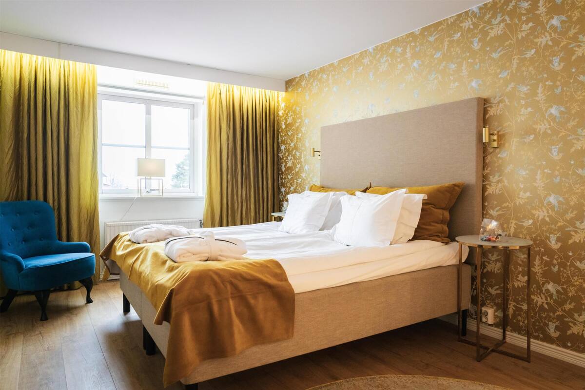 A hotel room with a bed, a blue armchair, a round side table, and a window with curtains.