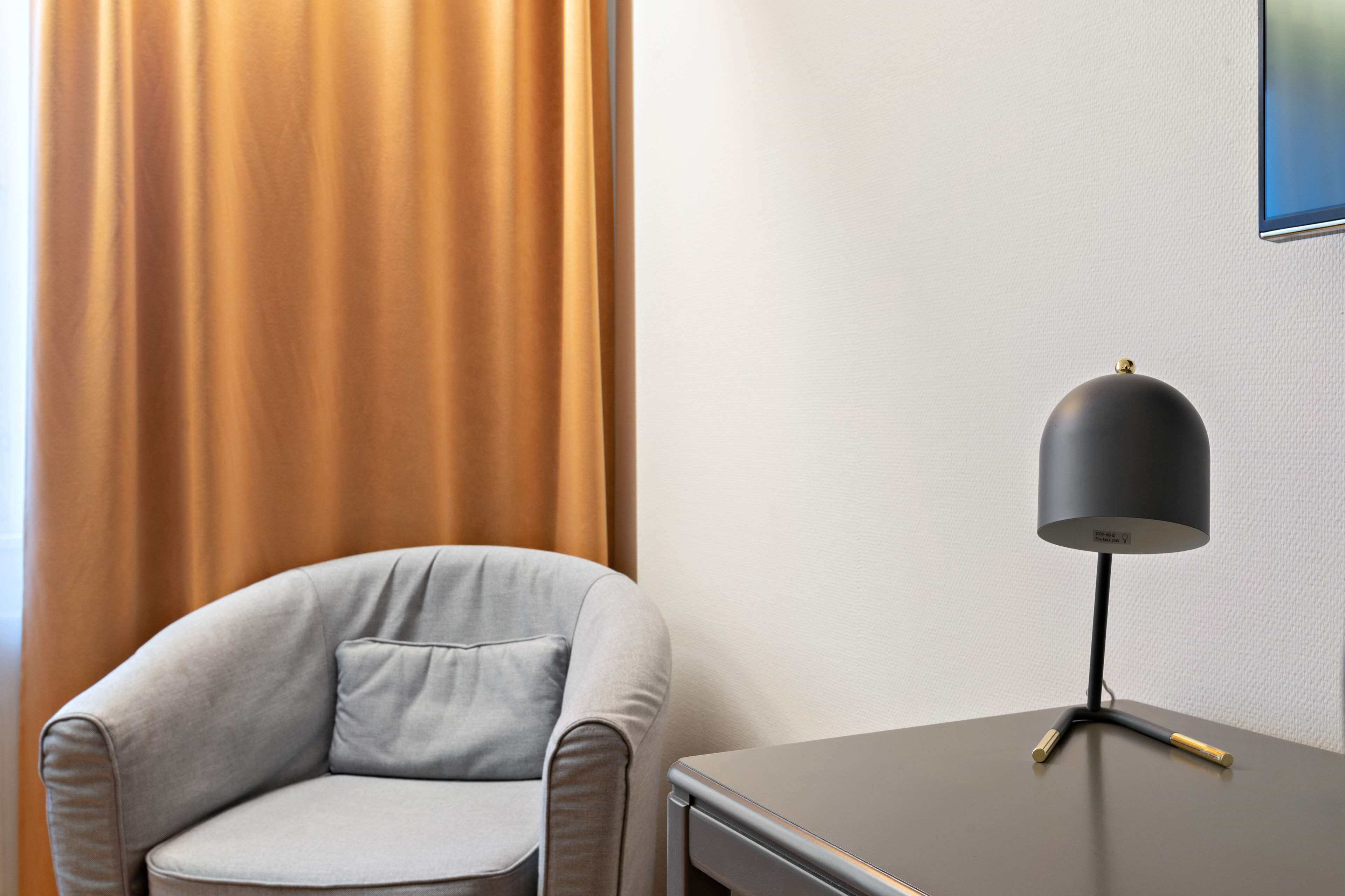 A corner of a hotel room with a grey armchair and a small table with a lamp.
