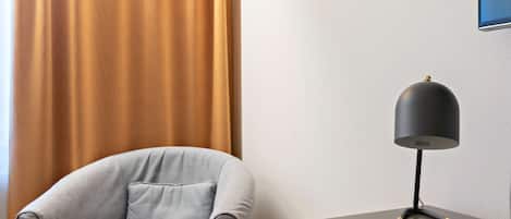 A corner of a hotel room with a grey armchair and a small table with a lamp.
