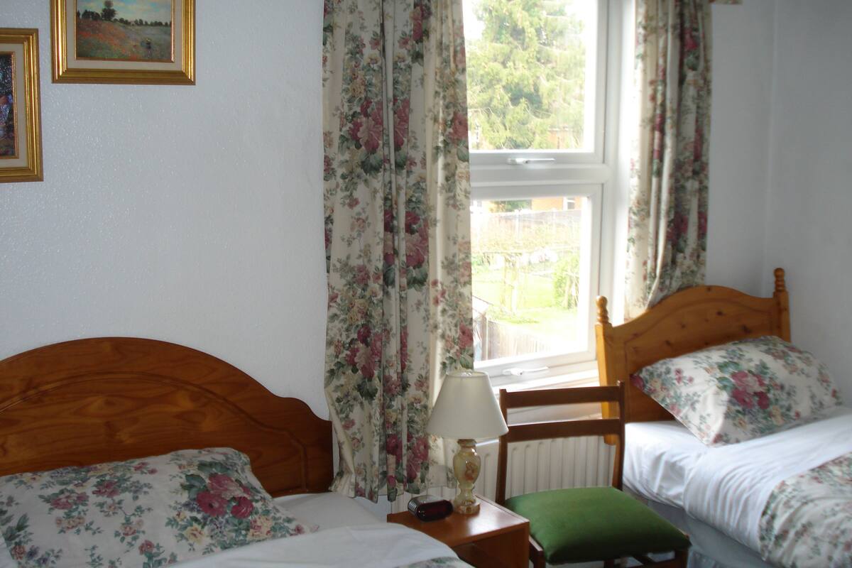 A bedroom with two single beds, floral curtains, a wooden headboard, a bedside table with a lamp, and a window with a view of greenery.