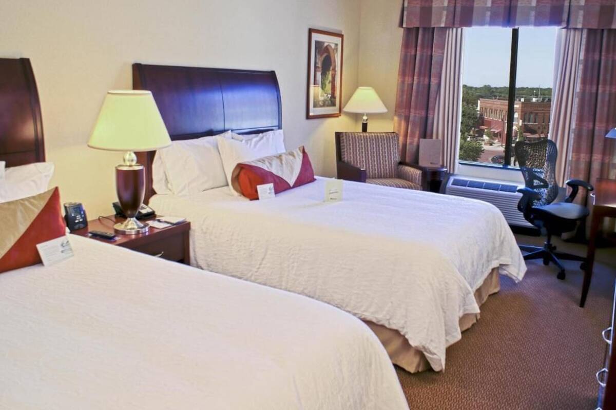 A hotel room with two beds, a desk, a chair, and a window with curtains.