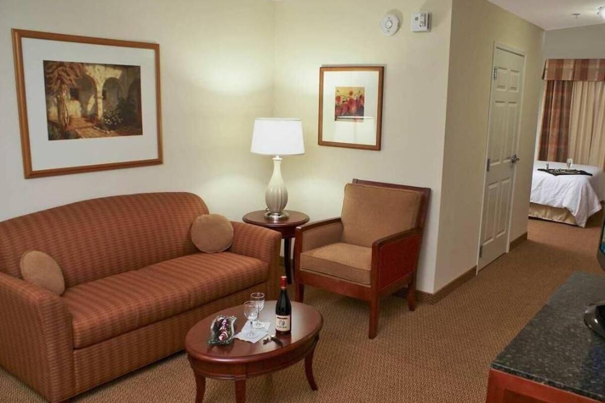 A hotel room with a sofa, armchair, small round table, lamp, and artwork on the walls.