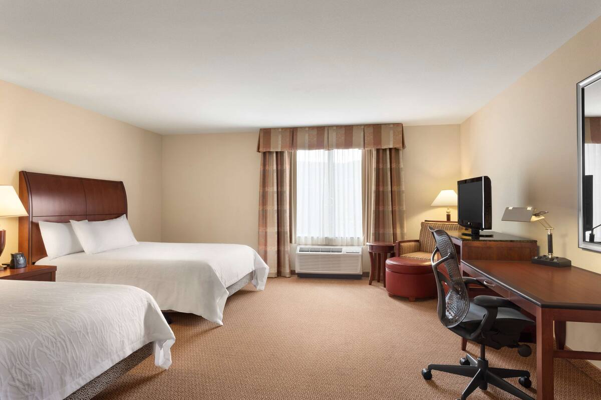 A hotel room with two beds, a desk with a chair, a television, and a window with curtains.