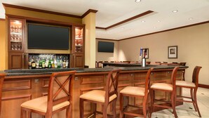 A bar area with wooden bar stools, a countertop with bottles, and a television mounted above the bar. - Hilton Garden Inn Bartlesville (Bartlesville)