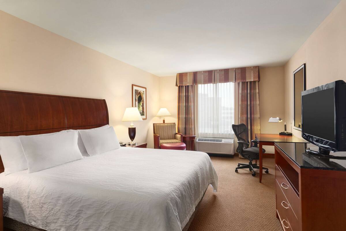 A hotel room with a large bed, a desk with a television, an air conditioning unit, and a window with curtains.