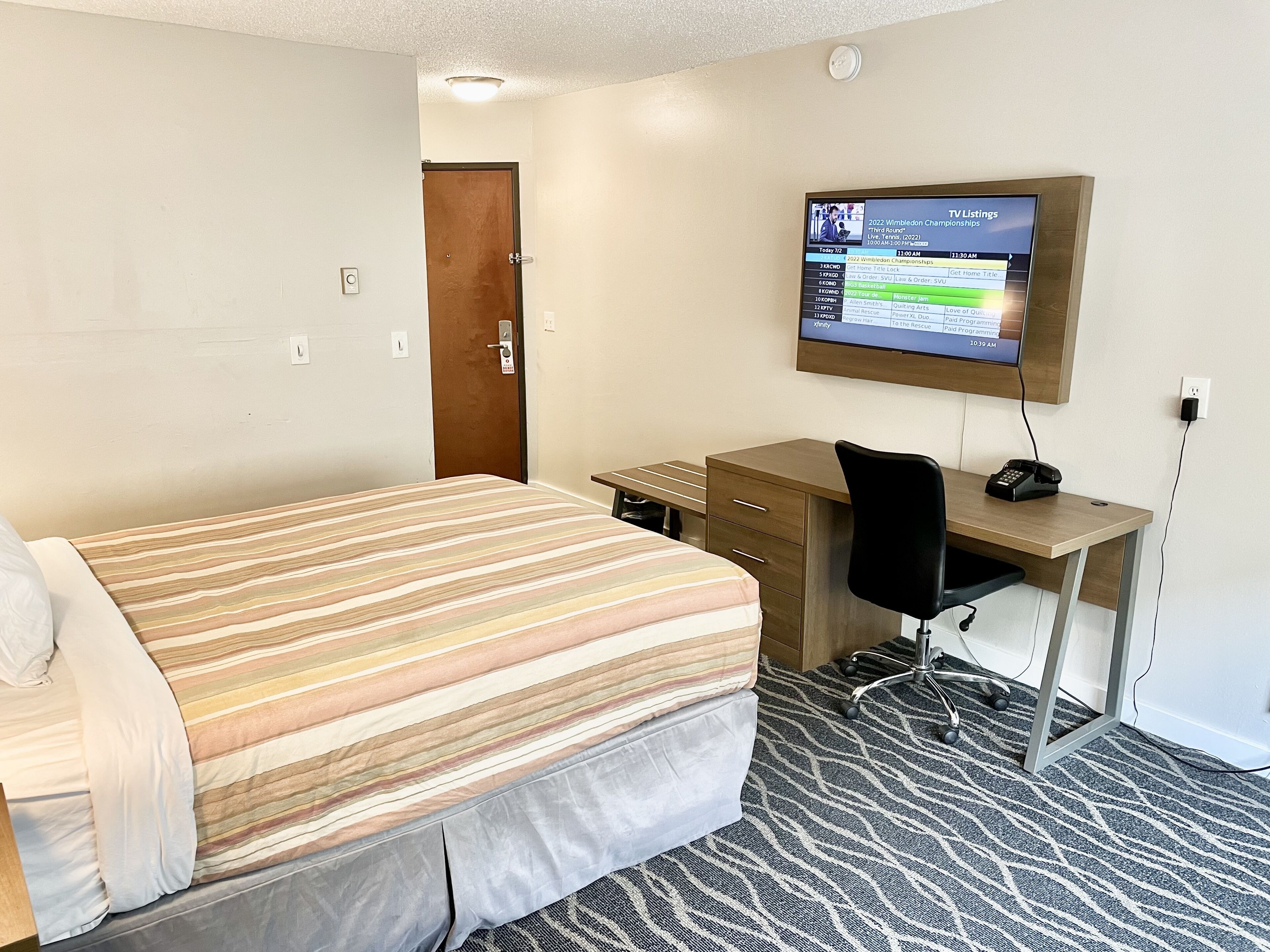 A hotel room with a bed, a desk, a chair, a TV mounted on the wall, and a carpeted floor.