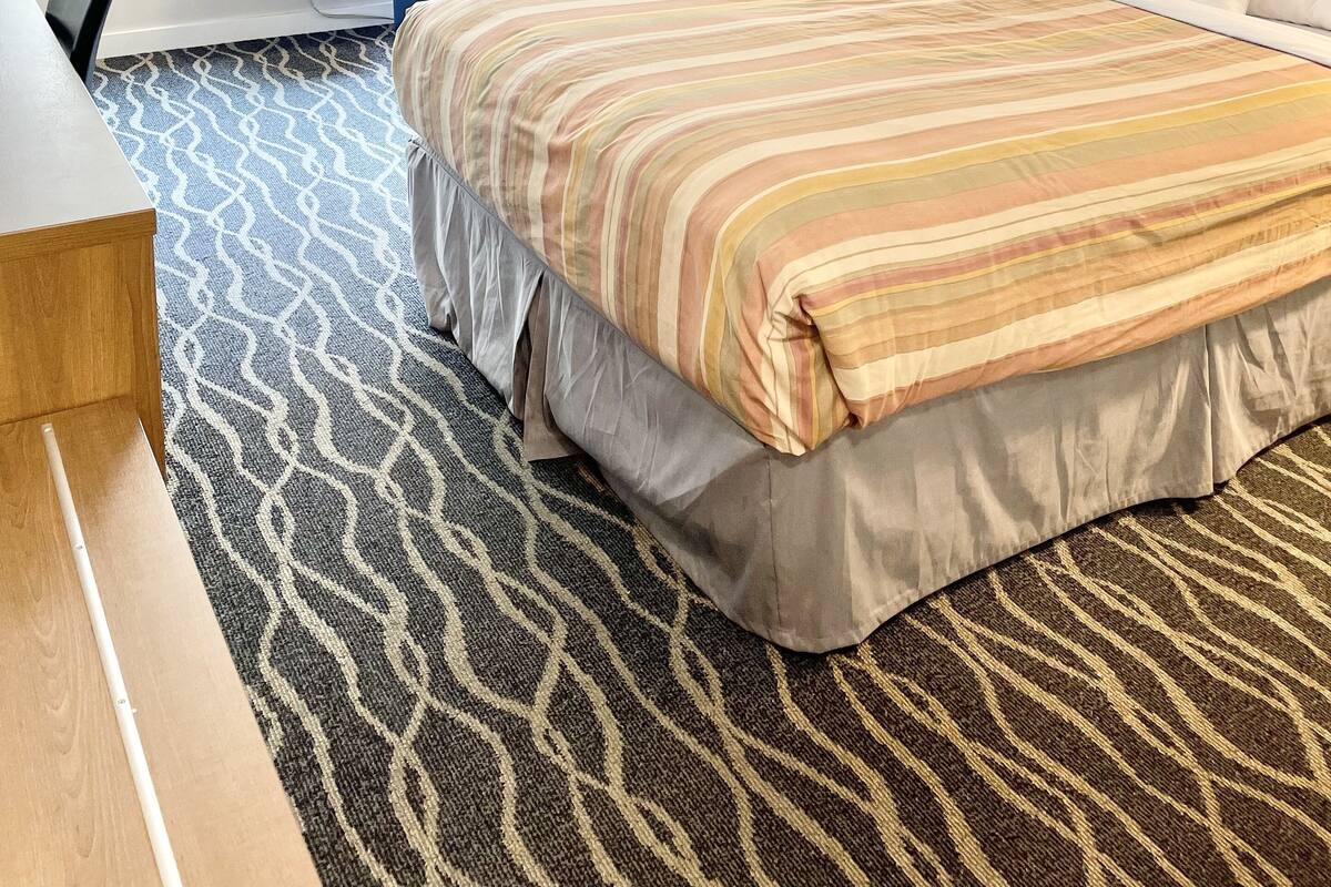 A hotel room with a large bed, a desk, a chair, a nightstand, and a window with blinds.