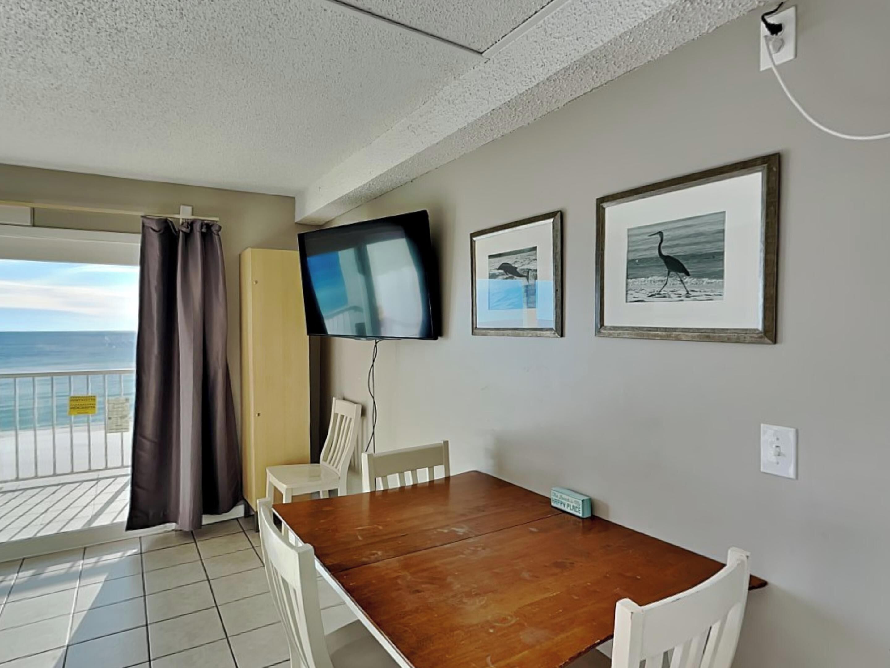 Una habitación con una mesa de madera y sillas blancas, un televisor montado en la pared y cuadros con imágenes de pájaros.
