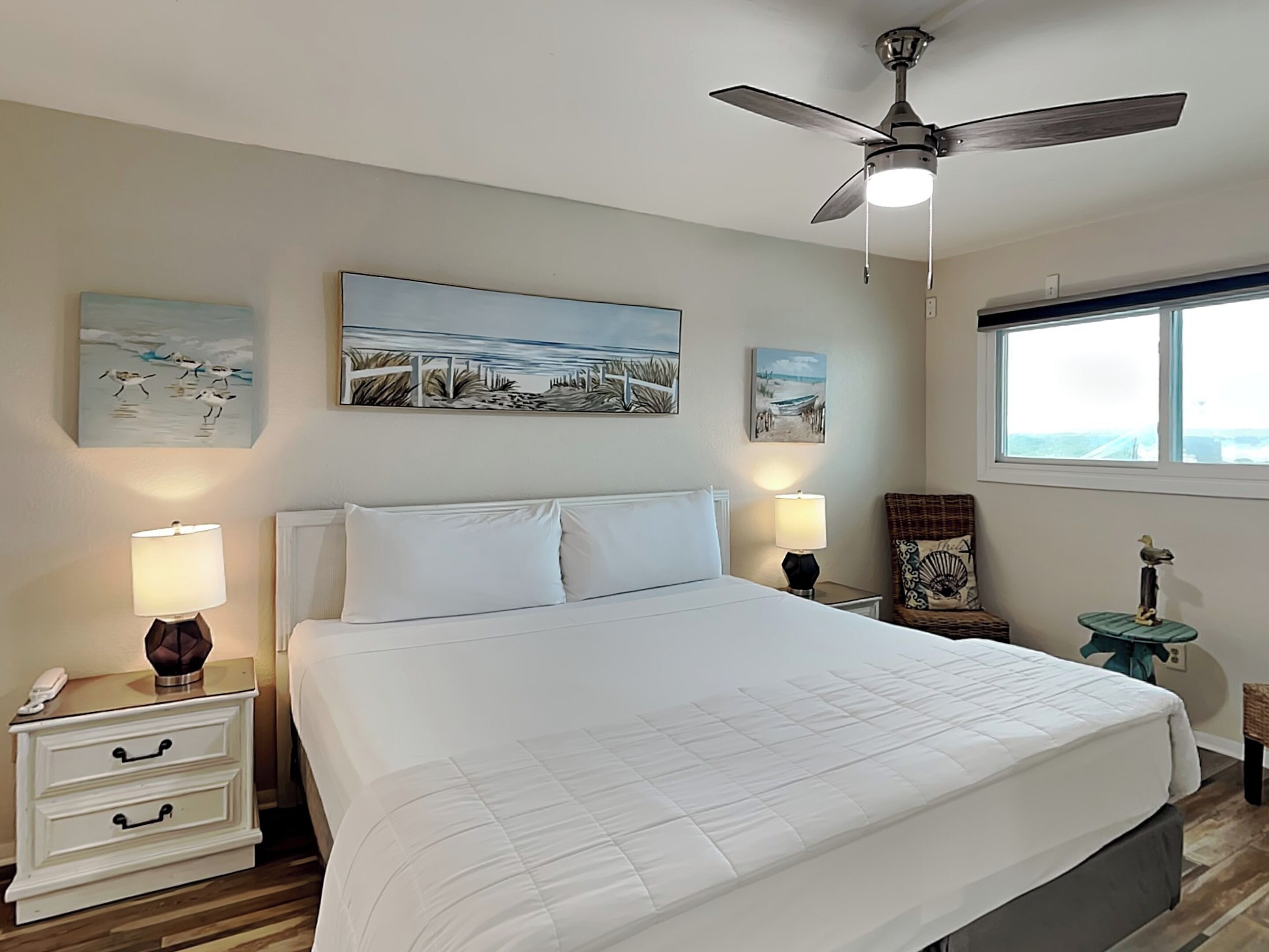 A bedroom with a bed, two bedside tables with lamps, a ceiling fan, and artwork on the walls.