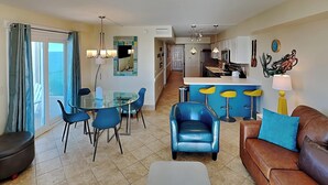 A modern living room with a glass dining table, blue chairs, a brown leather sofa, and a kitchen area with a bar. - Summit Beach Resort by Resort Collection (Panama City Beach)