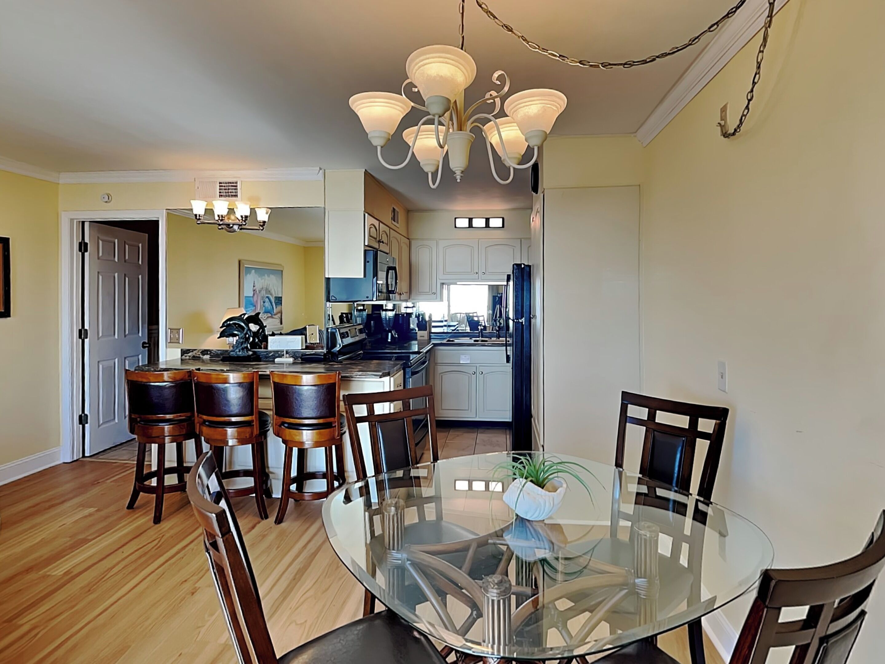 A dining area with a glass table and wooden chairs, a kitchen with white cabinets and a refrigerator, and a chandelier above.