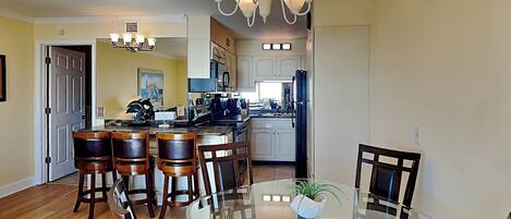 A dining area with a glass table and wooden chairs, a kitchen with white cabinets and a refrigerator, and a chandelier above.