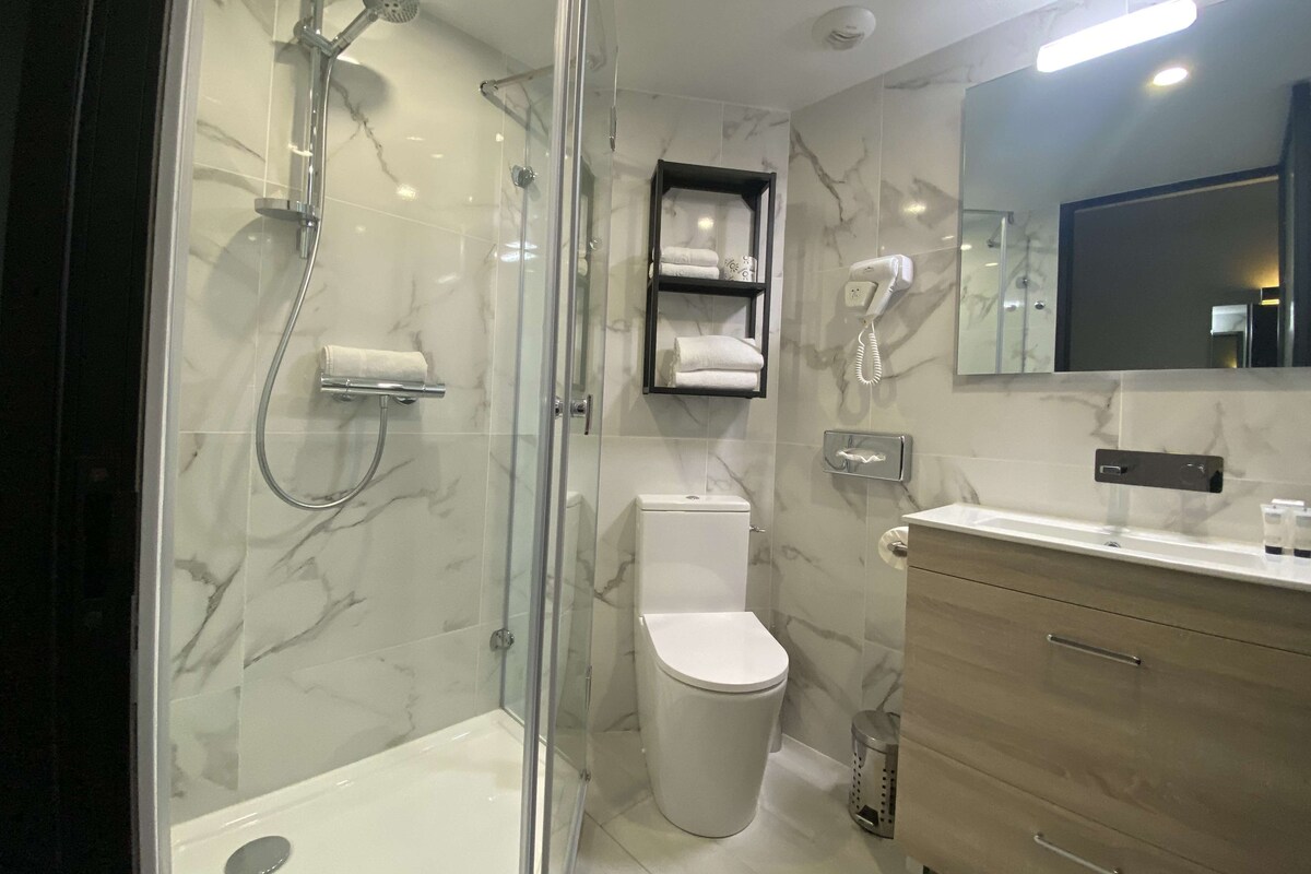 Un baño moderno con ducha envuelta en vidrio, inodoro, tocador con espejo y repisa con toallas.