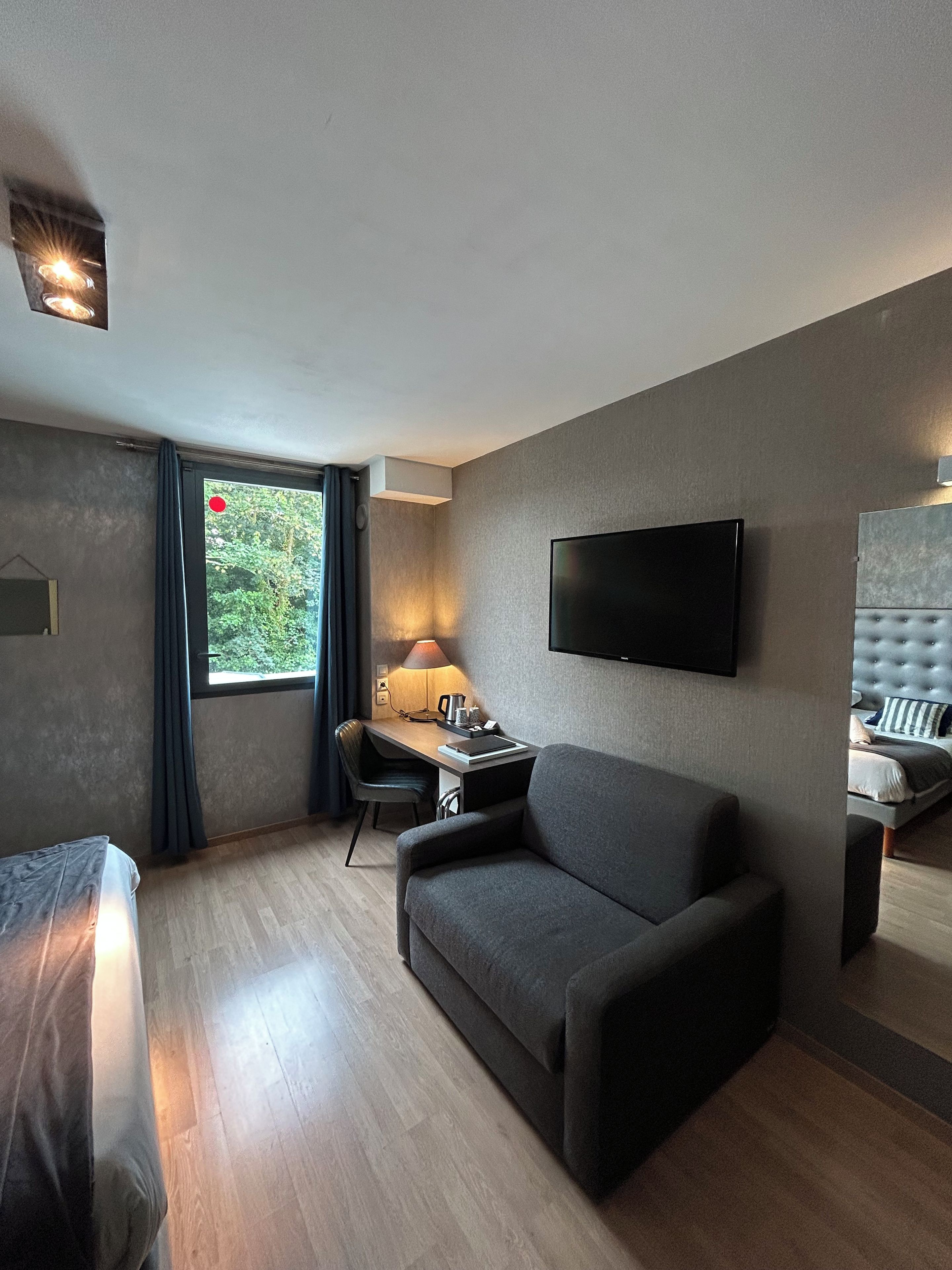 Habitación de hotel con cama, sofá, escritorio con lámpara, televisor montado en la pared y ventana con cortinas.