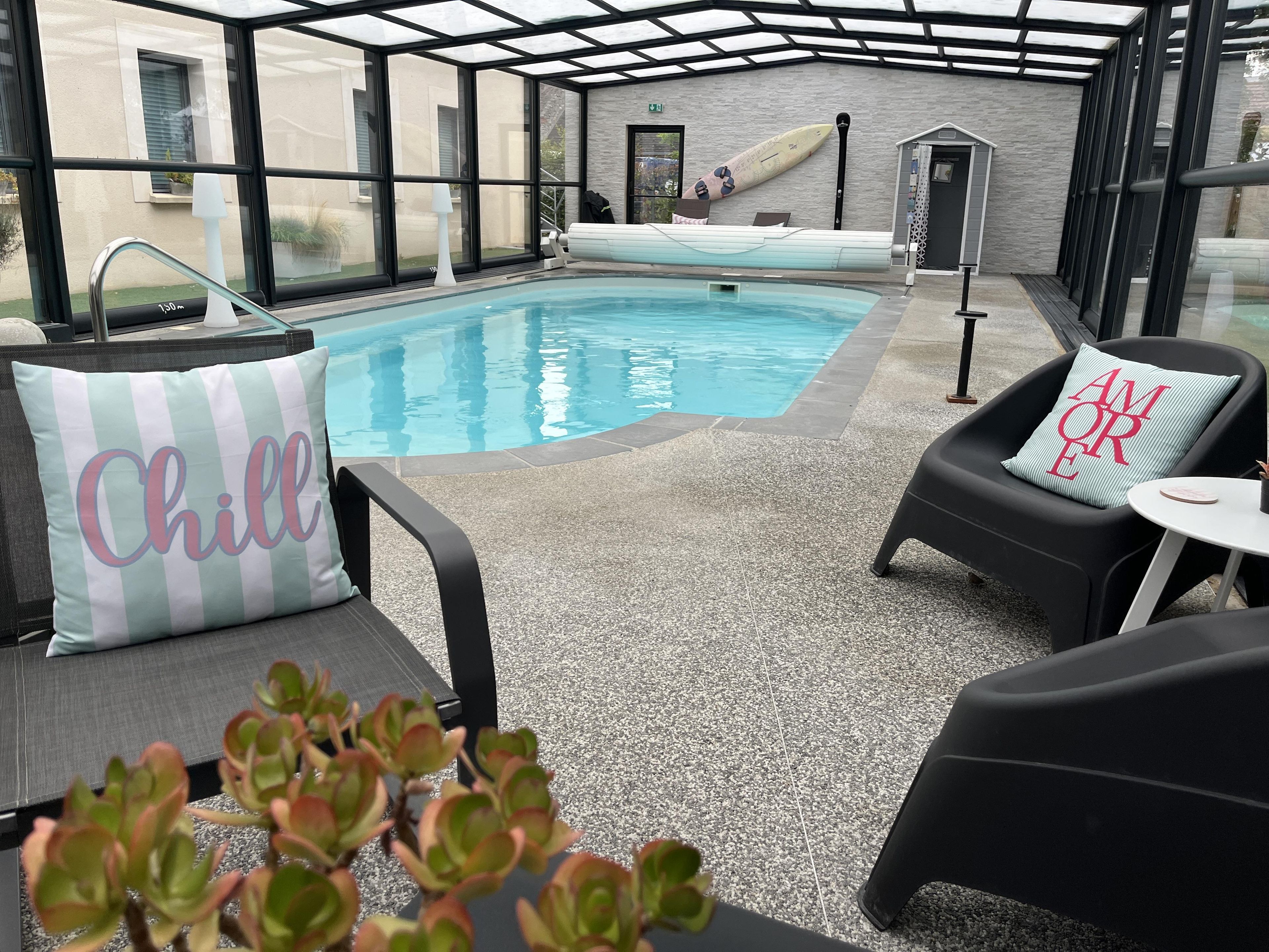 Un espace piscine intérieure avec des chaises longues, une petite table et un coussin orné du mot «Chill».