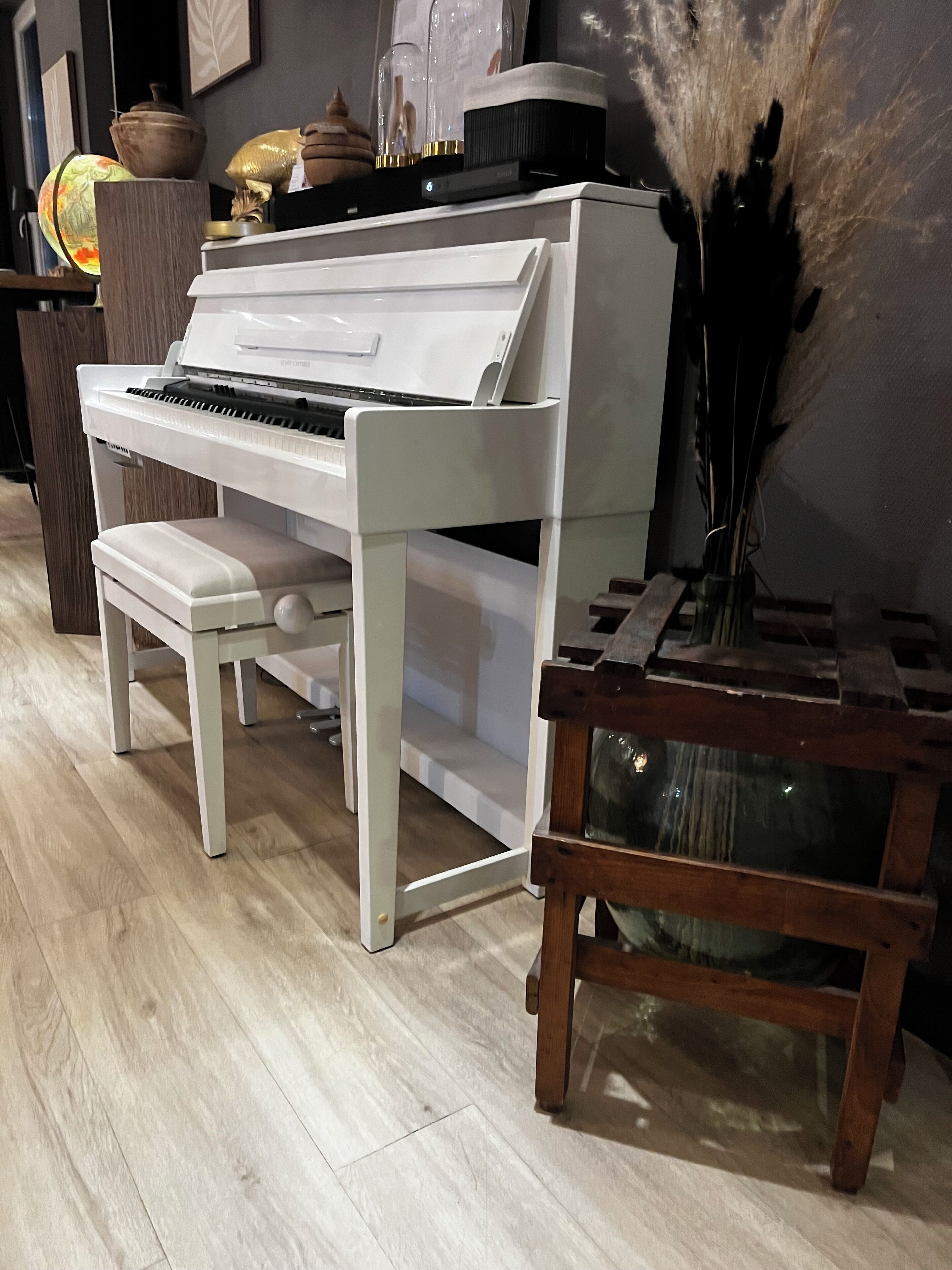 A white piano with a bench, a wooden side table with a vase, and decorative items on a shelf.