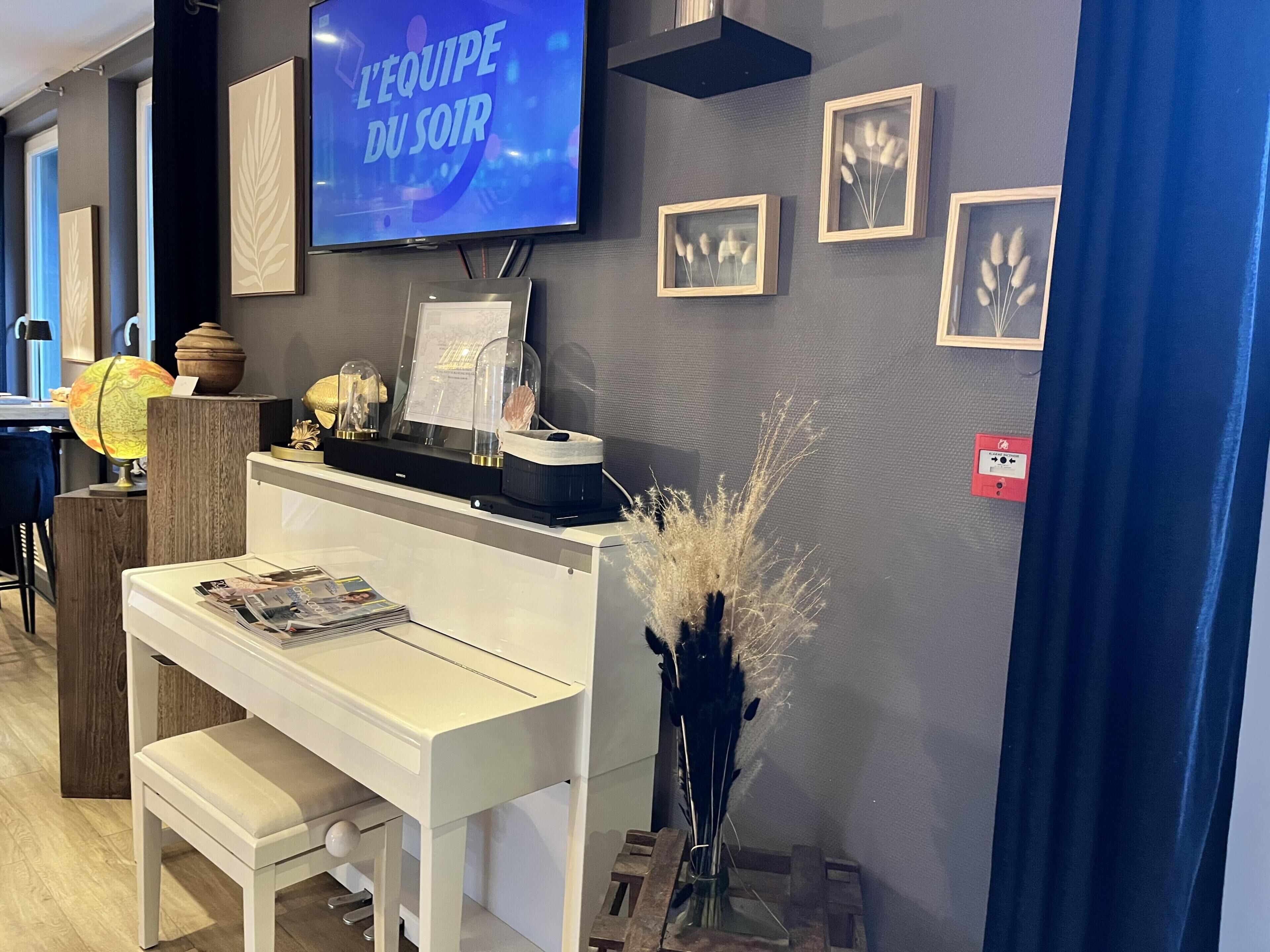 Una habitación con una televisión mostrando 'L'EQUIPE DU SOIR', un escritorio blanco con una banca, un globo terráqueo, objetos decorativos y cuadros en la pared.