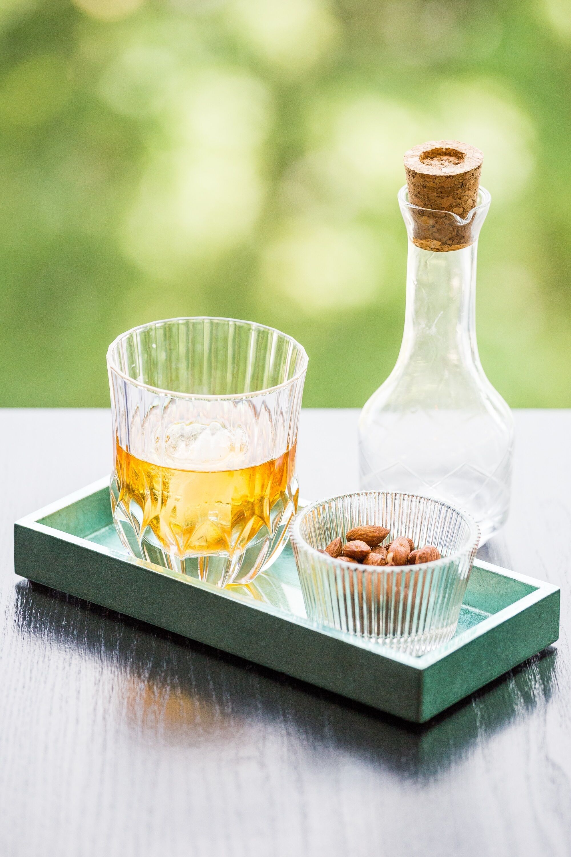 Un verre contenant un liquide ambré, posé sur un plateau vert, une bouteille transparente bouchée d’un bouchon et un petit bol d’amandes.