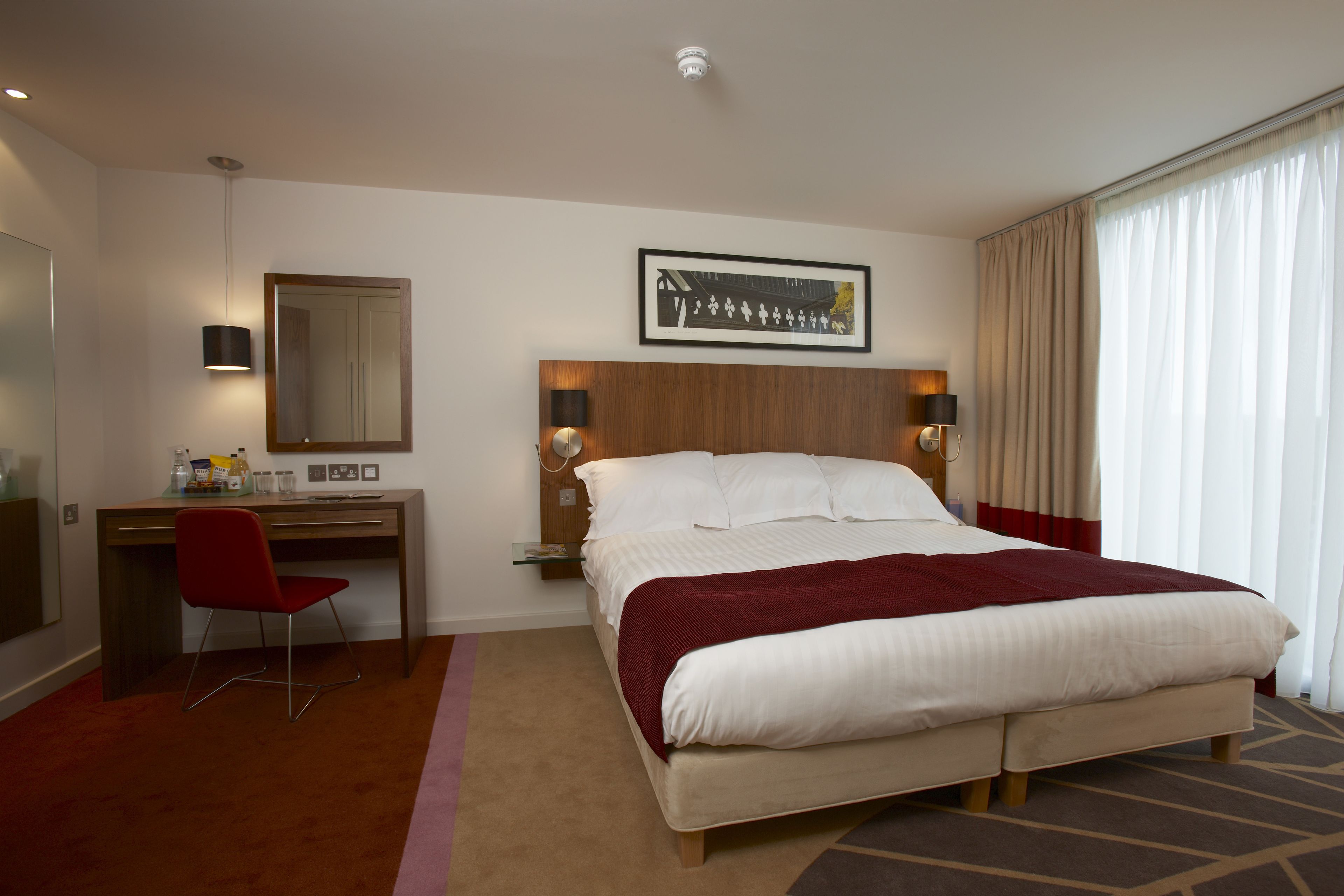 Une chambre d’hôtel avec un lit, un bureau, une chaise, un miroir, un tableau encadré et une fenêtre avec des rideaux.