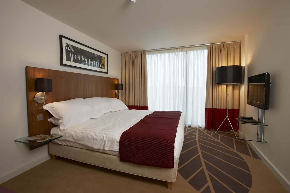 A hotel room with a large bed, a bedside table with a lamp, a TV, and a framed picture on the wall.