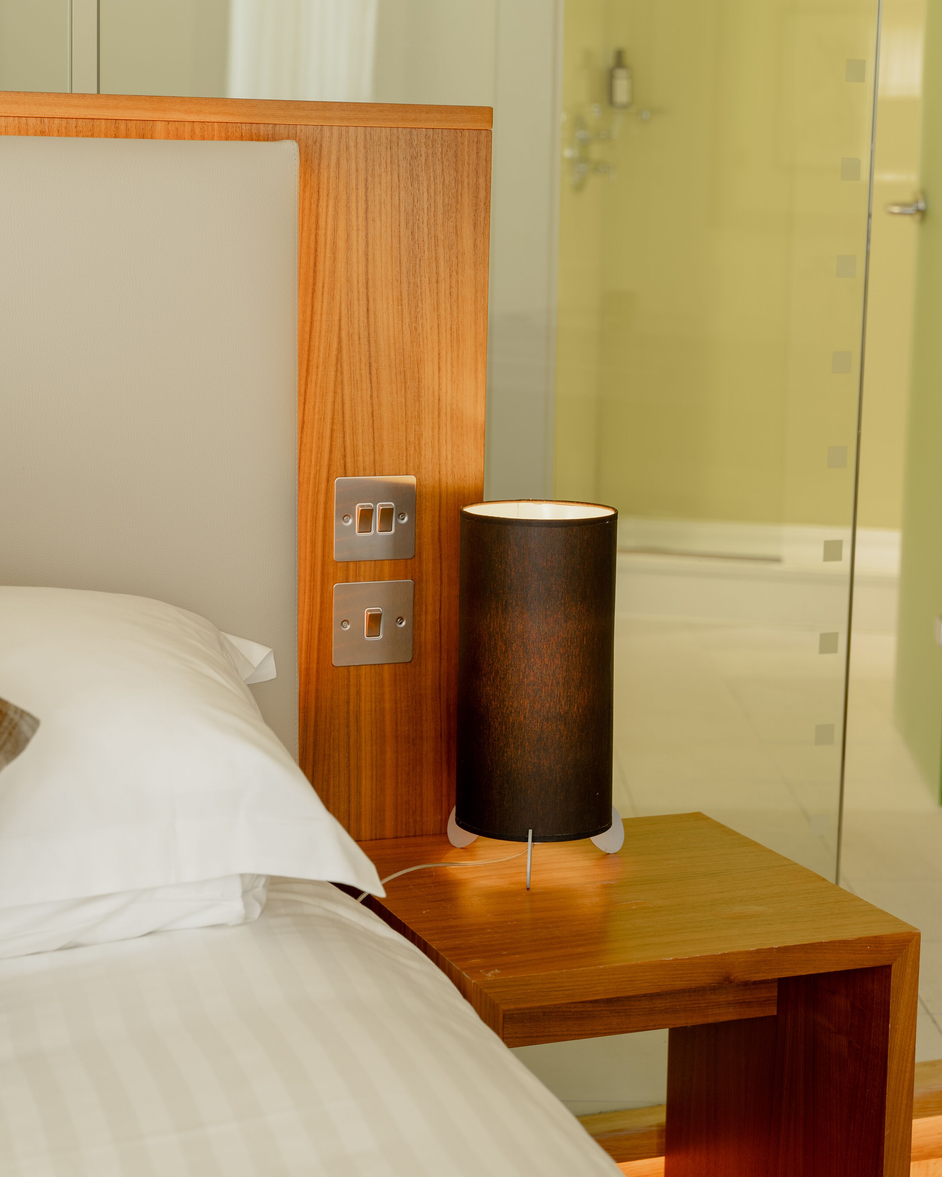 A neatly made bed with white linens, a wooden nightstand with a lamp, and a wall-mounted light switch.
