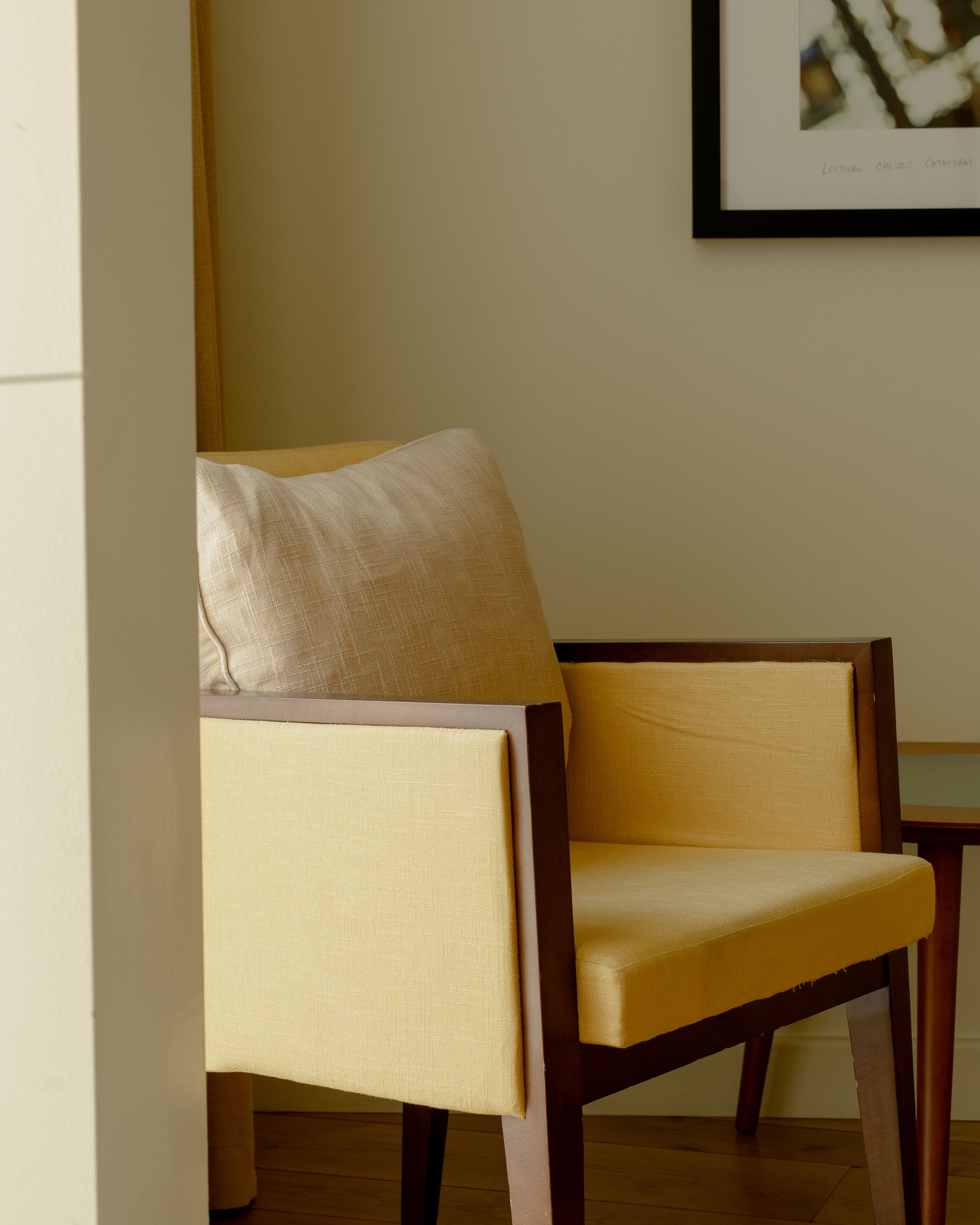 A chair with a cushion and a wooden frame, positioned in a room with a wooden floor and a framed picture on the wall.