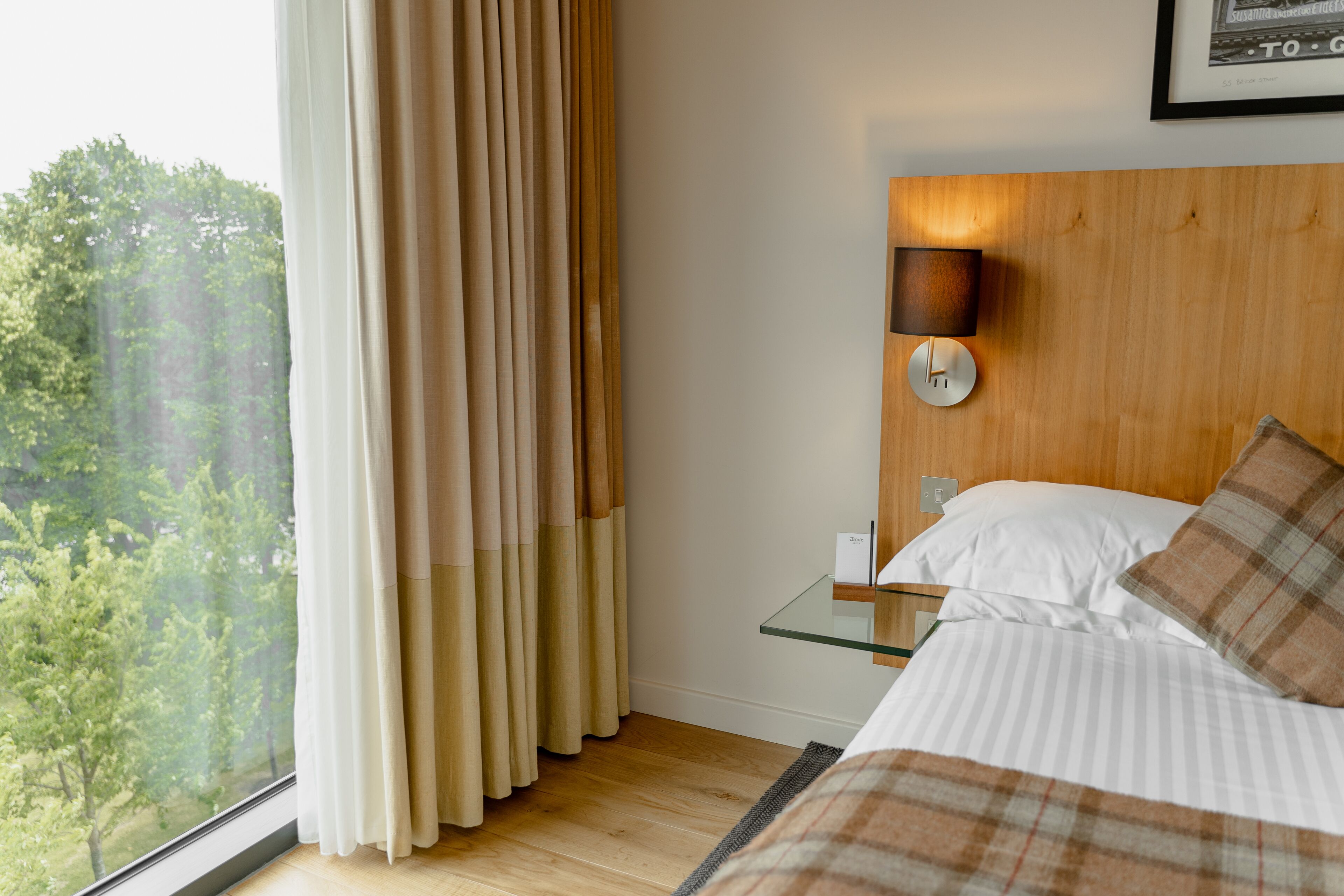 Een hotelkamer met een bed, nachtkastje, een wandlamp en een raam met gordijnen.