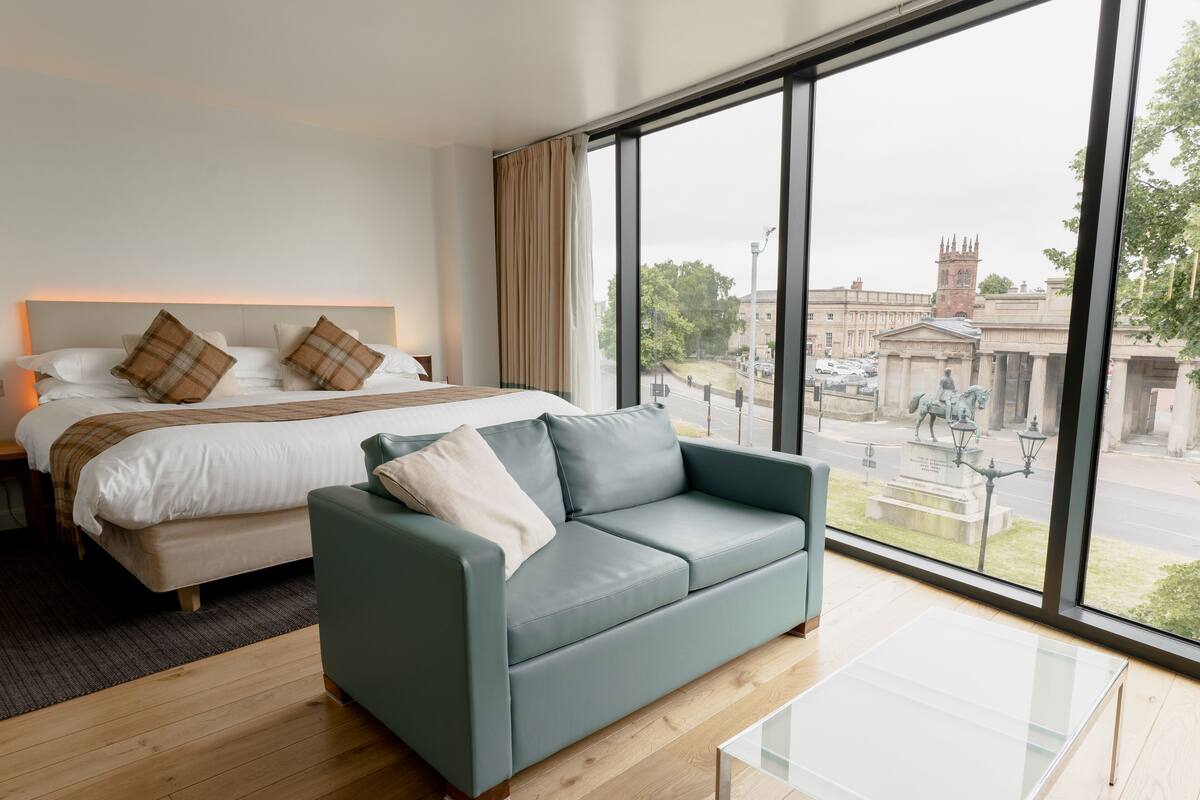 A modern hotel room with a large bed, a grey sofa, and a glass coffee table.