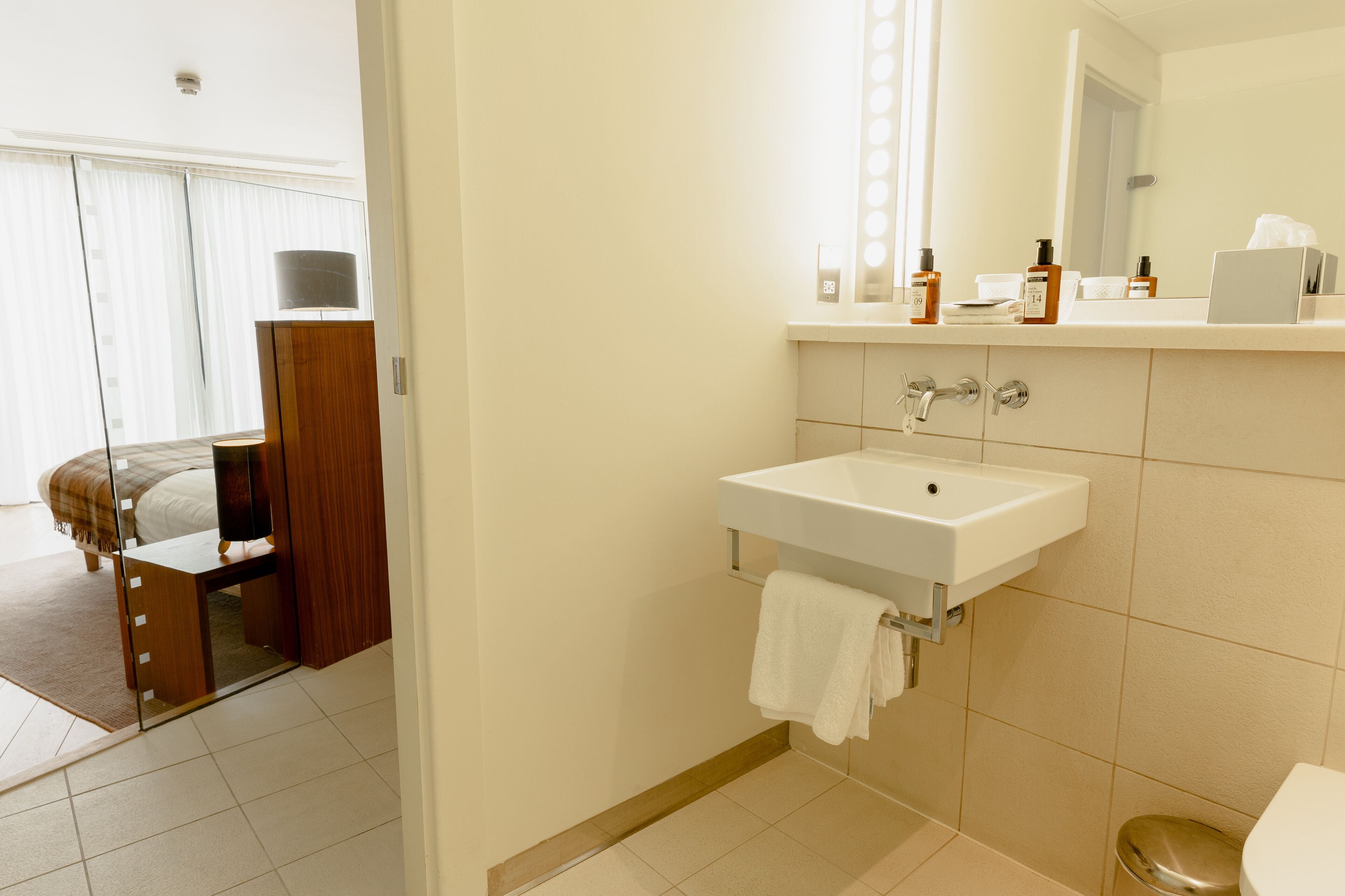 A modern bathroom with a white sink, mirror, and toilet. There is a towel hanging on the sink and toiletries on the countertop.