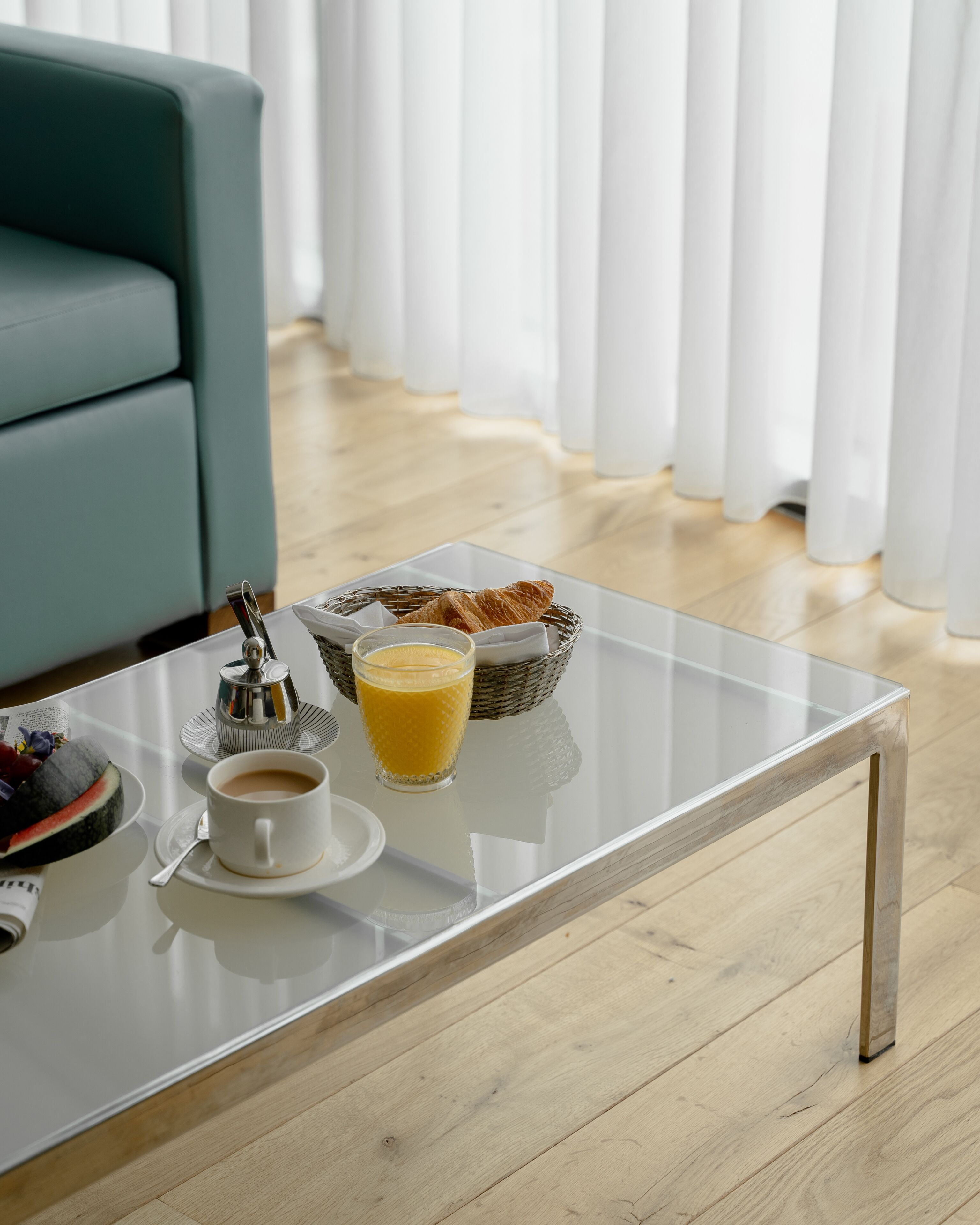 Un salon moderne avec une table basse en verre, un panier de croissants, un verre de jus d’orange et une tasse de café.