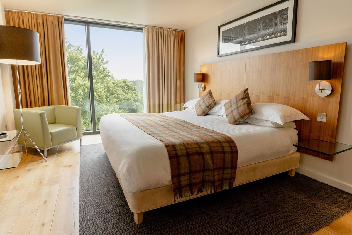 A modern hotel room with a large bed, a sitting area with a chair, a glass side table, and a view of the outdoors.