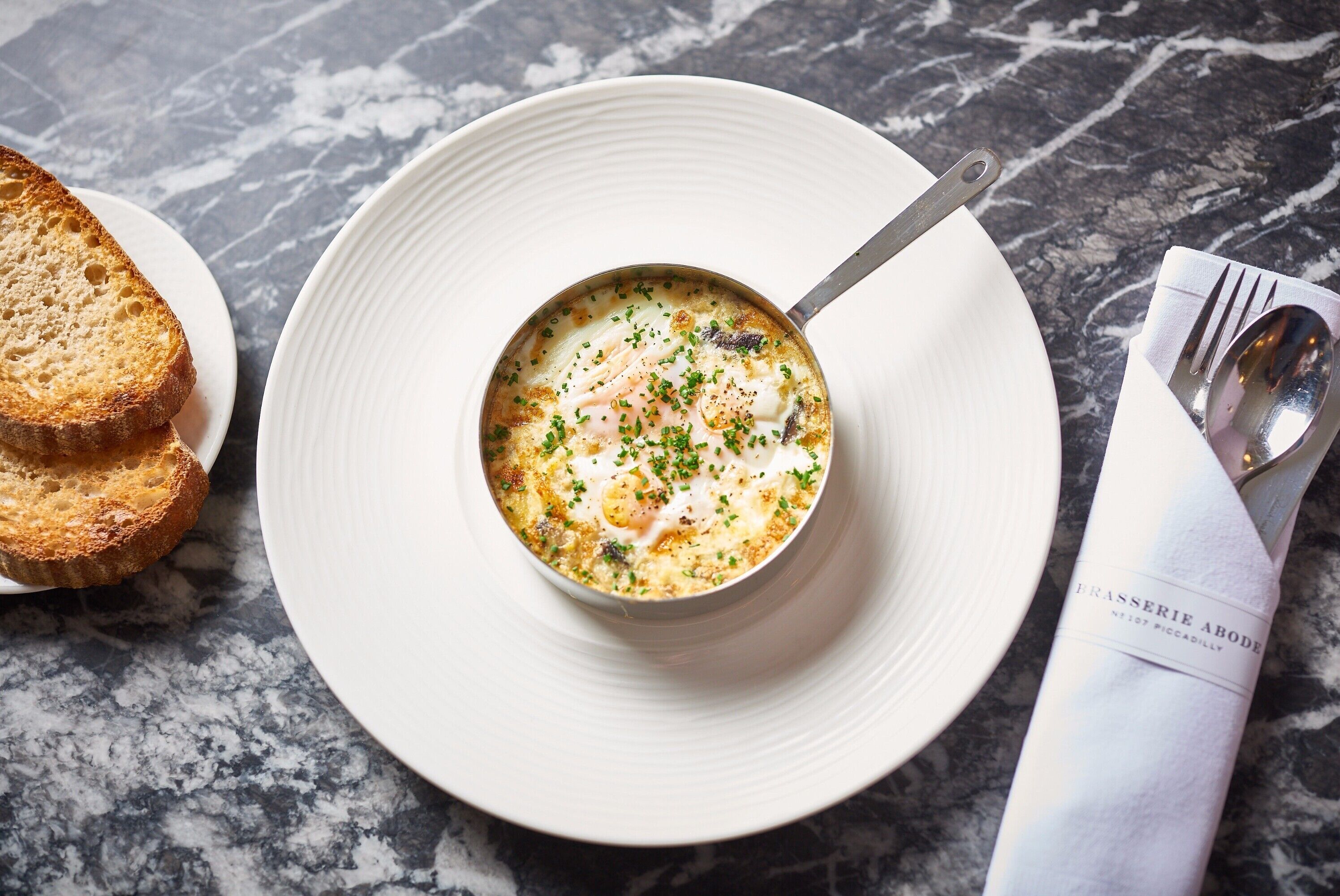 Un bol de soupe sur une assiette blanche, accompagné d’une cuillère, ainsi que de deux tranches de pain grillé et d’une serviette sur laquelle repose une cuillère.