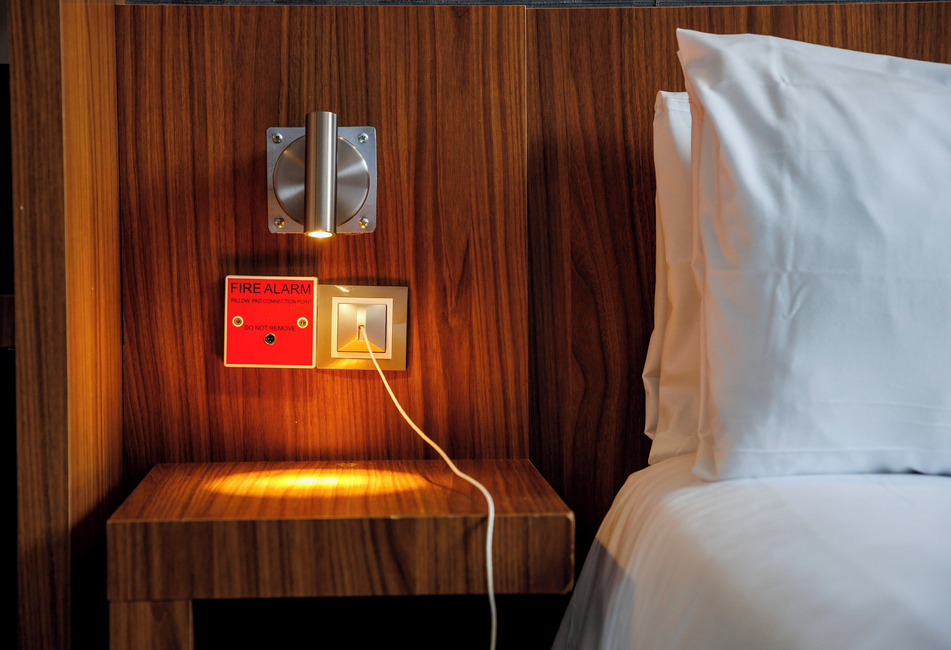 A wooden bedside table with a fire alarm and a light switch.