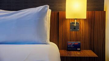 A neatly made bed with white linens and a single pillow, a wall-mounted lamp, and a sign on a wooden nightstand that reads "FIRM OR SOFT IT'S UP TO YOU!".