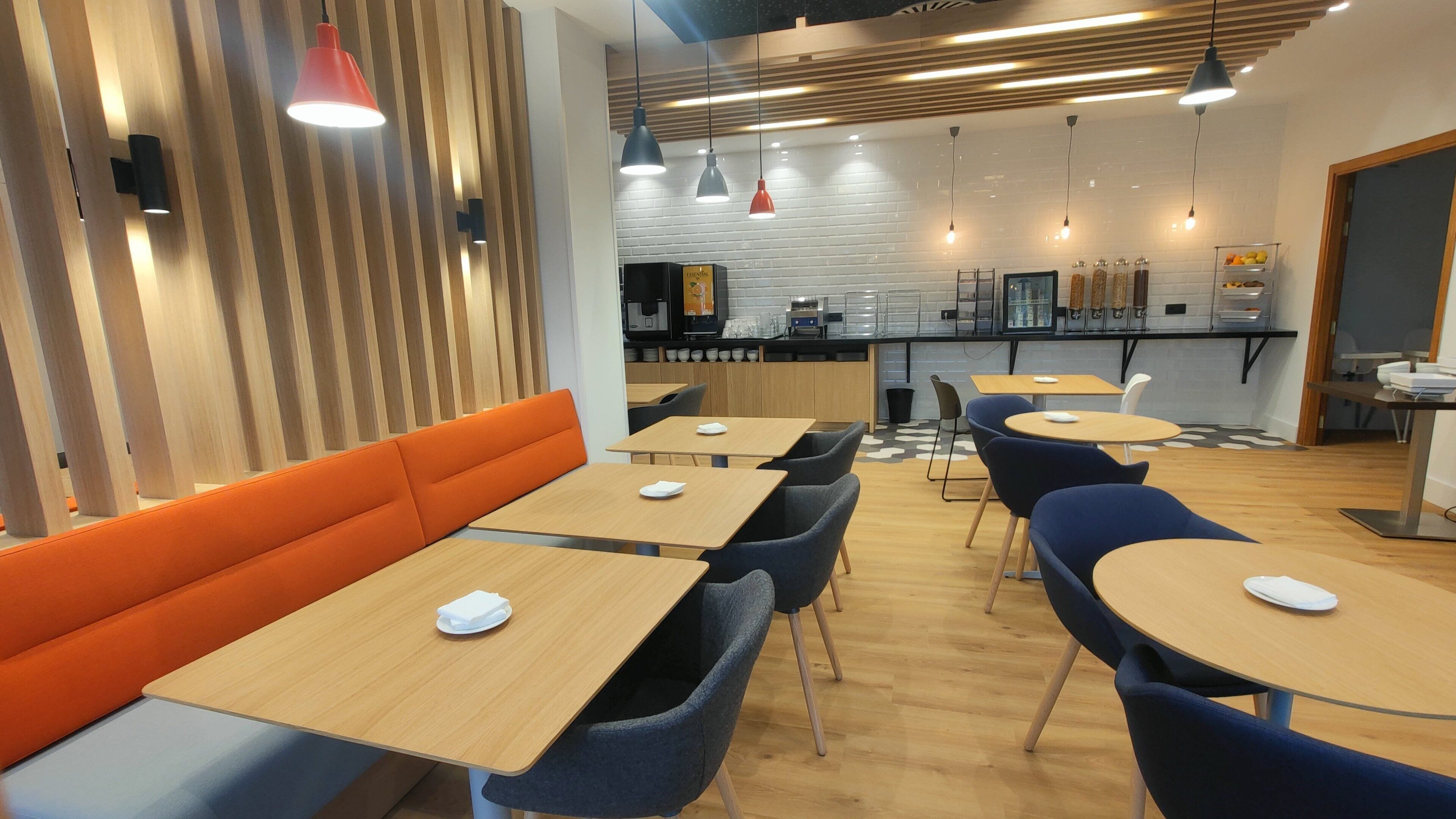 Un comedor moderno con asientos de madera y tapicería, una cocina al fondo e iluminación colgante.