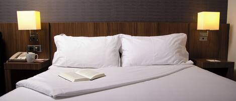 A neatly made bed with white linens, two bedside lamps, a telephone, and a book on the nightstand.