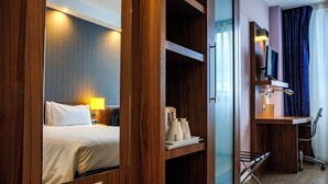 Habitación de hotel con cama, mesita de noche, lámpara, televisor, escritorio, silla y una pequeña estantería con objetos.