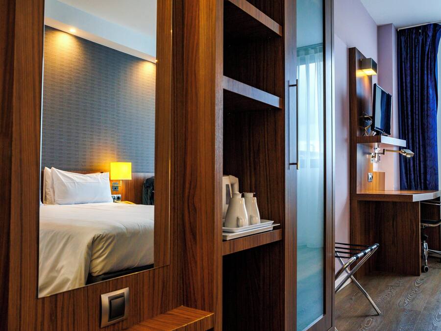 A hotel room with a bed, bedside table, lamp, TV, desk, chair, and a small shelf with items.