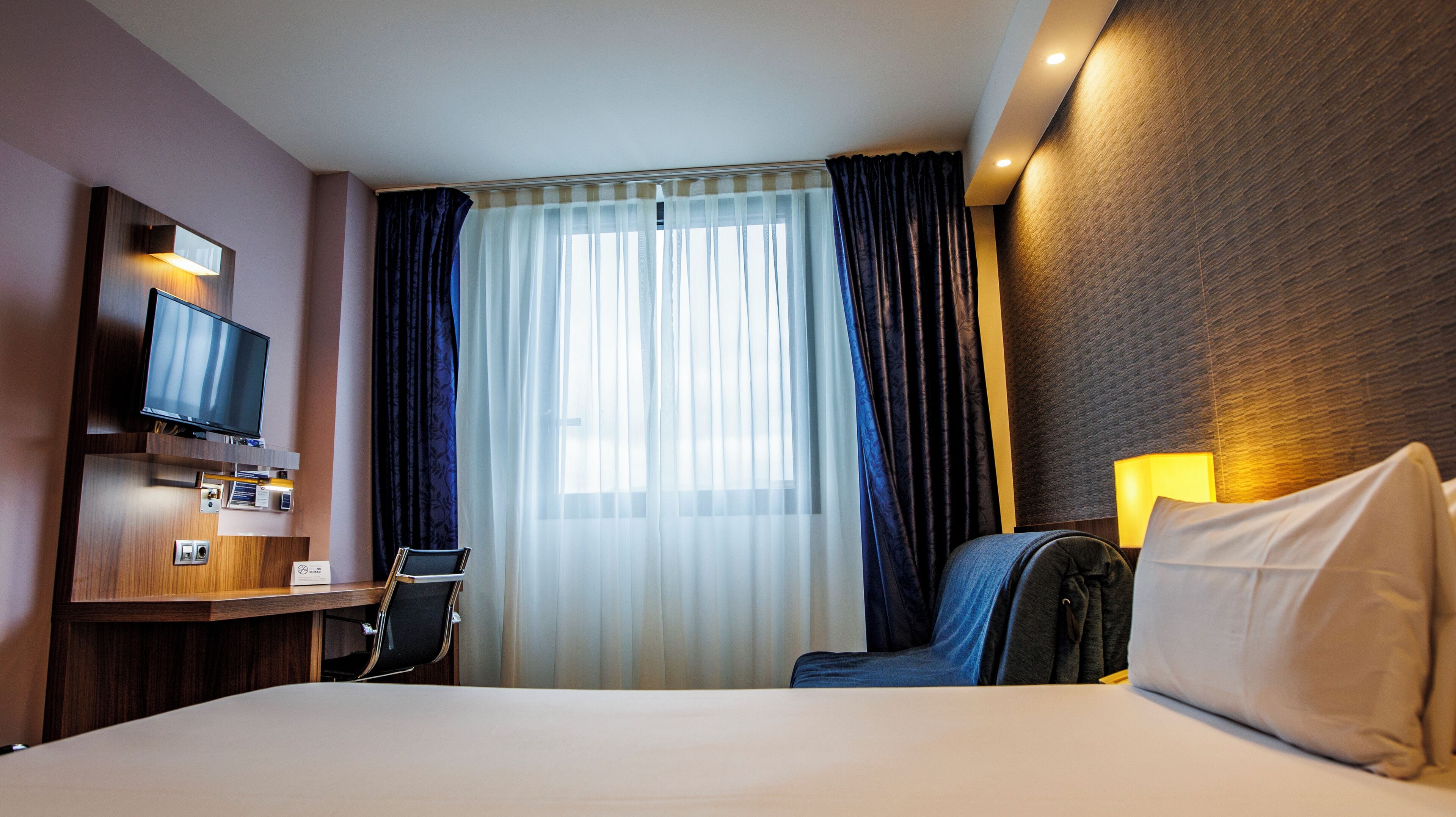 Habitación de hotel con cama, escritorio, silla, televisor y ventana con cortinas.