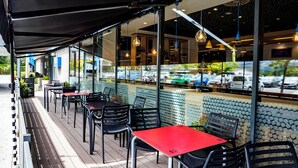 Un café al aire libre con mesas y sillas, una zona cubierta para sentarse y vistas a coches aparcados y vegetación.