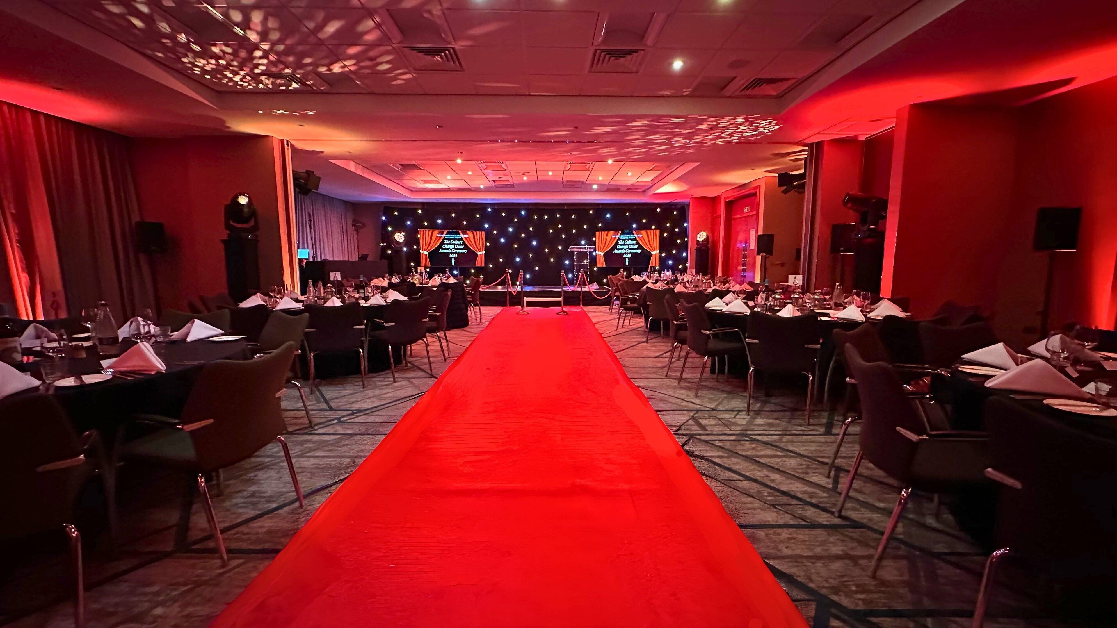 Un salón de banquetes con alfombra roja, mesas dispuestas para cenar y un escenario con cortinas.