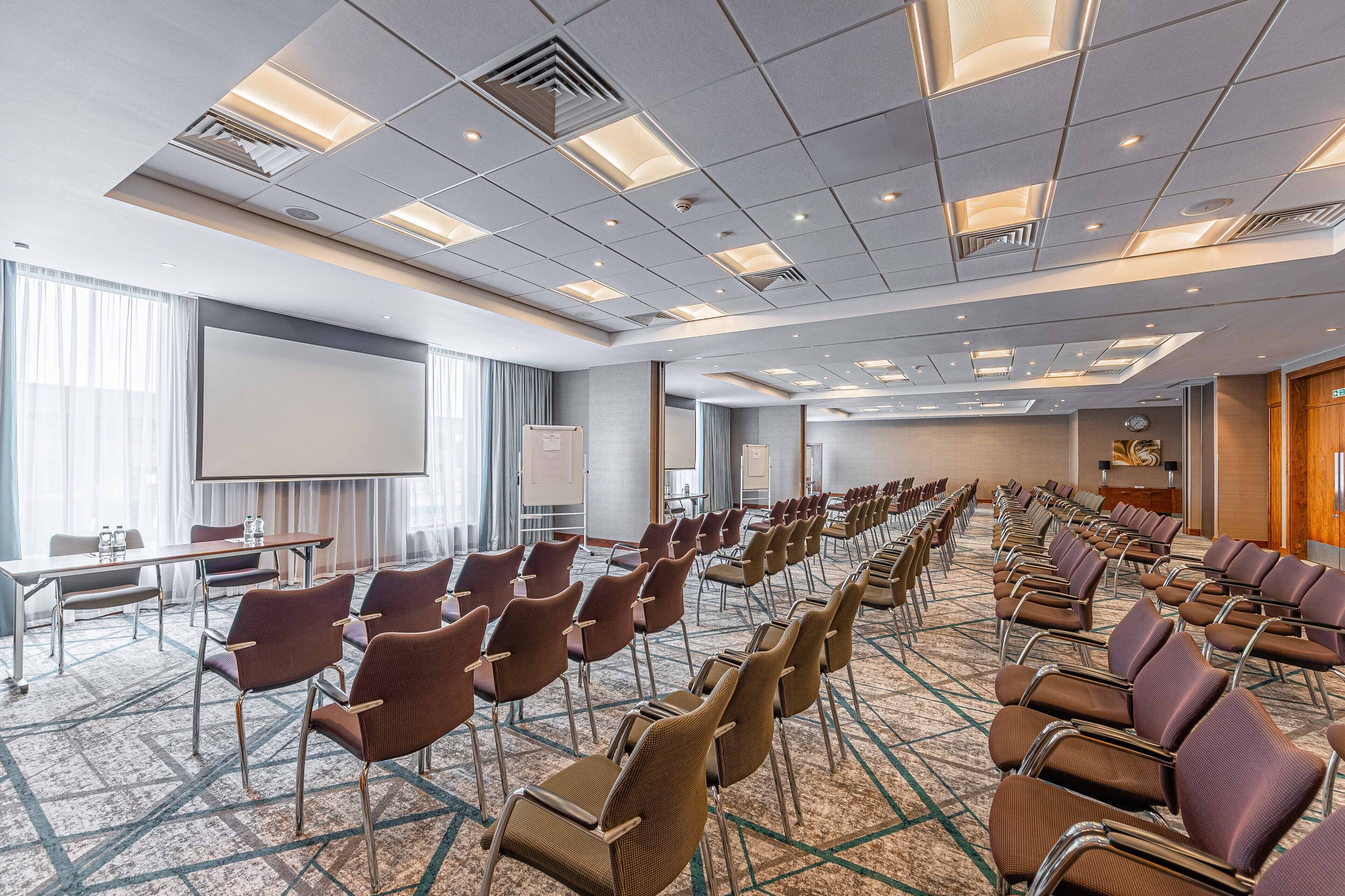 A conference room with rows of chairs, a projector screen, and a presentation area.