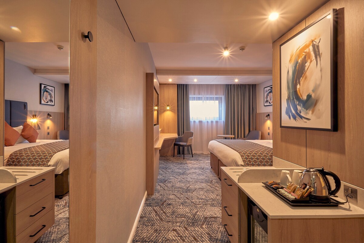 A hotel room with two beds, a desk, a chair, a bathroom, and a large painting on the wall.