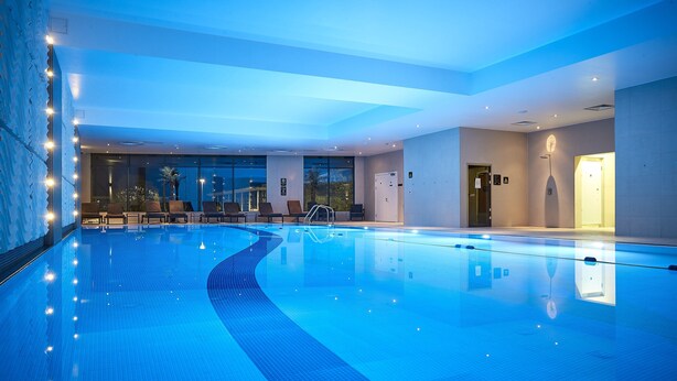 A modern indoor swimming pool with clear blue water, lounge chairs, and a view of the outdoors through large windows. - Crowne Plaza Reading by IHG (Reading)