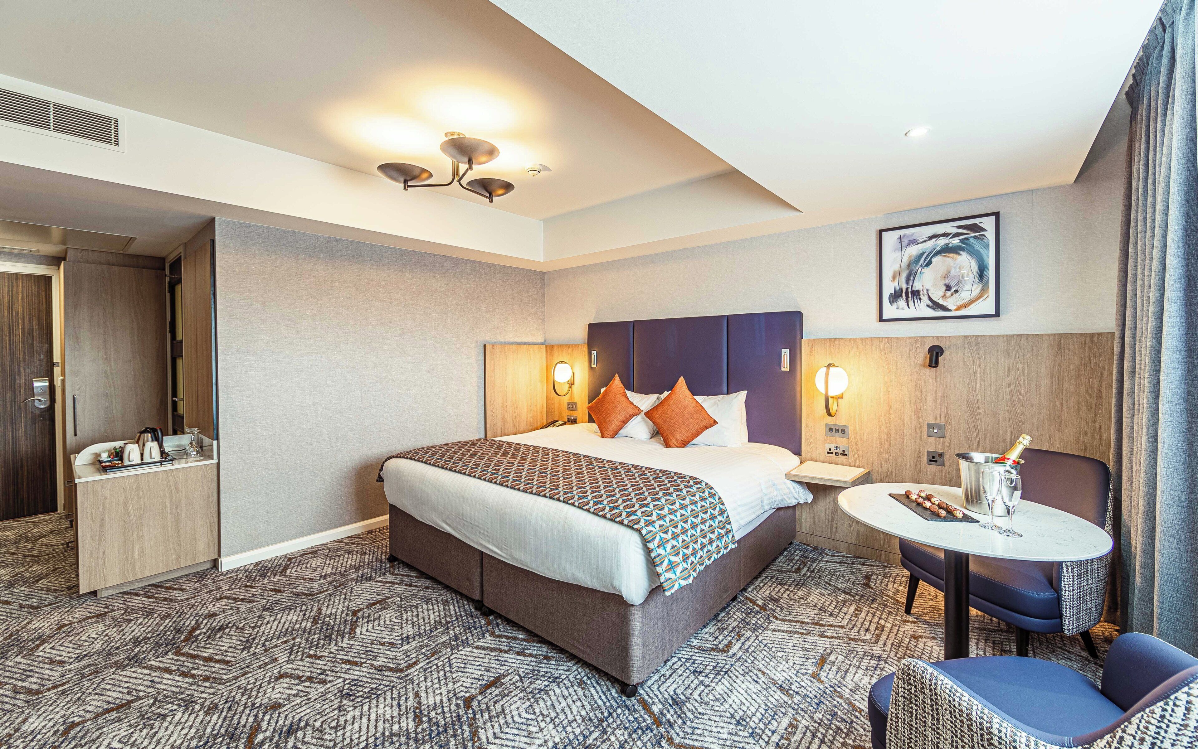 A hotel room with a large bed, bedside tables, a seating area with a table and chairs, and a painting on the wall.