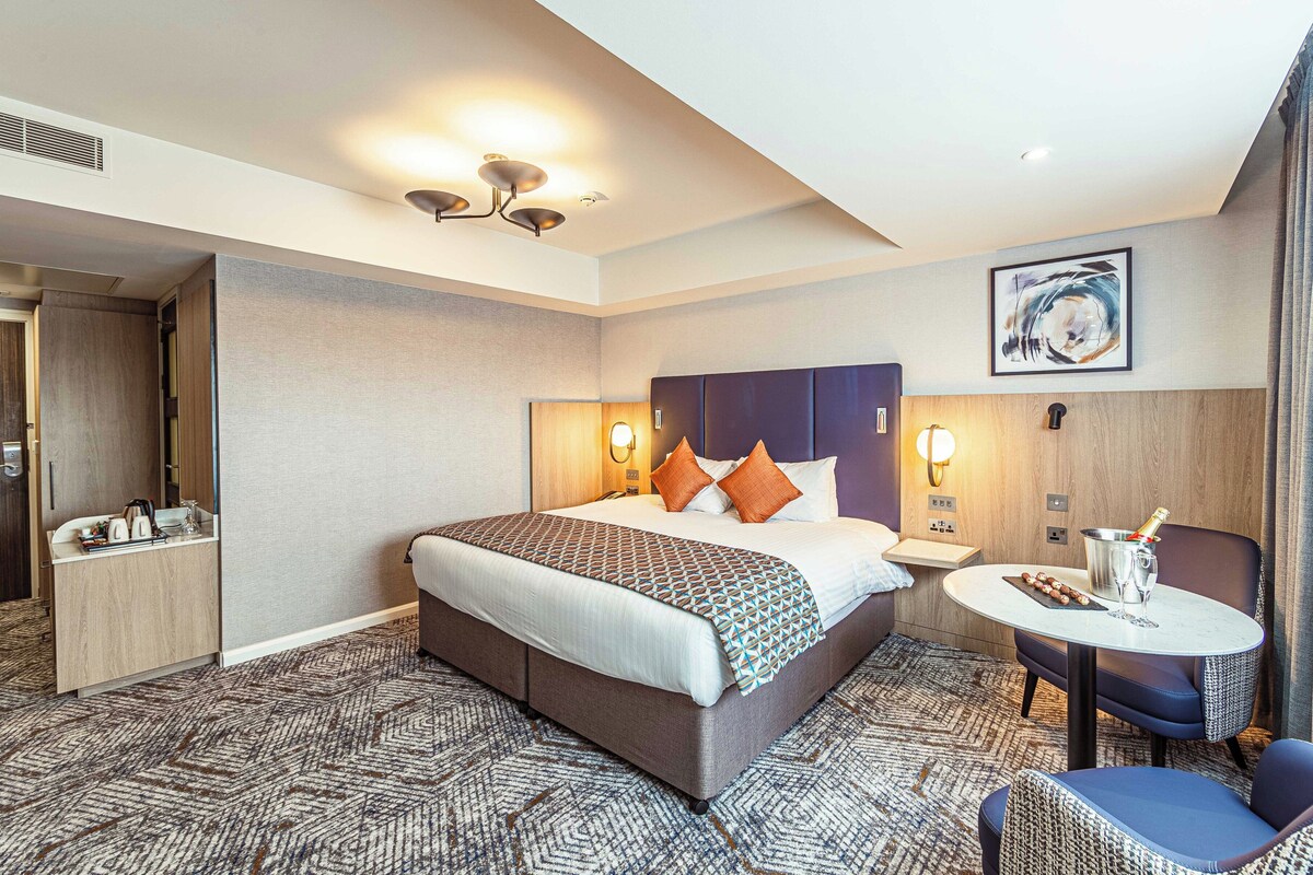 A hotel room with a large bed, bedside tables, a seating area with a table and chairs, and a painting on the wall.
