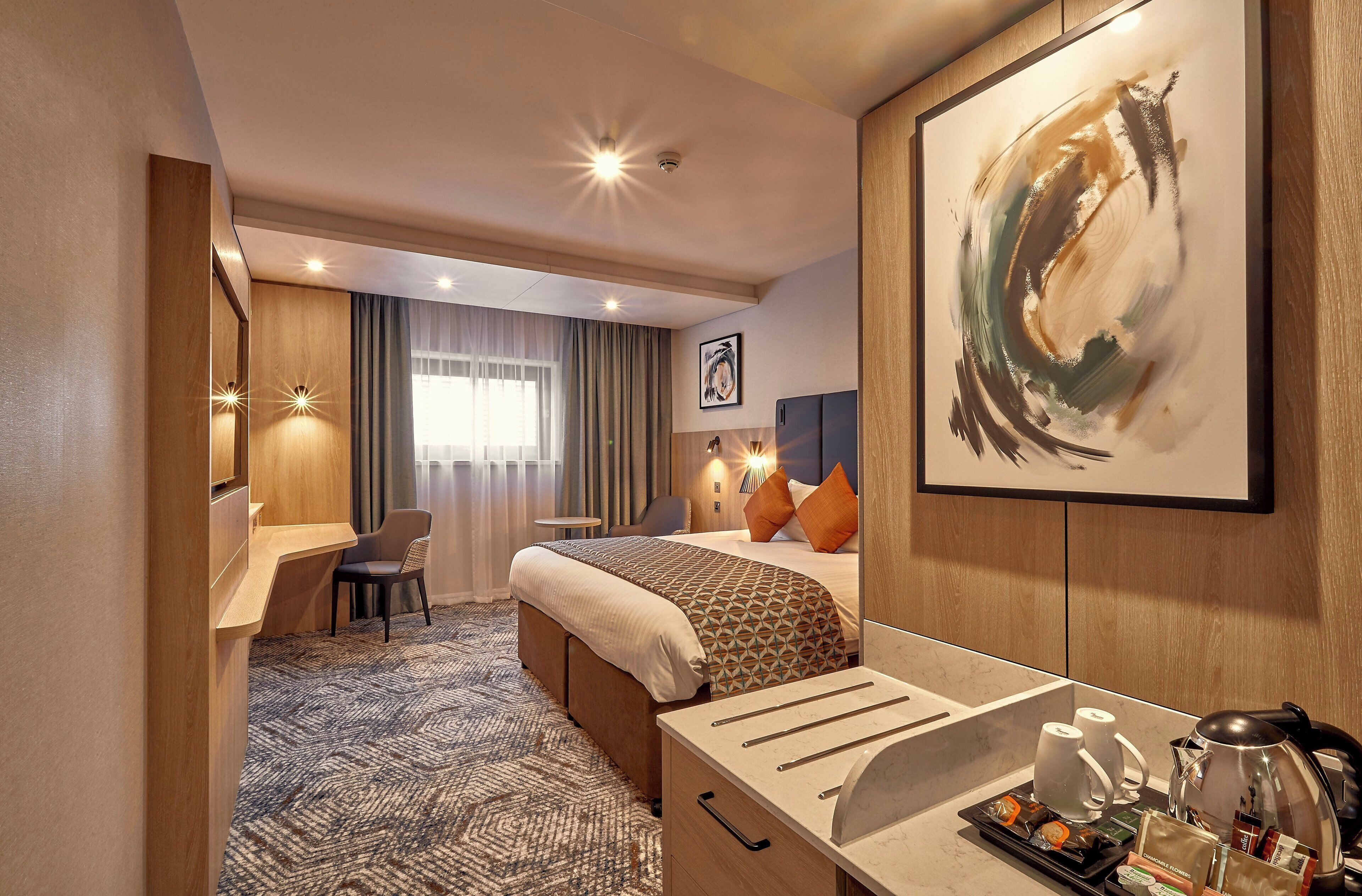 Een hotelkamer met een bed, een bureau, een stoel, een koffiezetapparaat en een lijst met abstracte kunst.