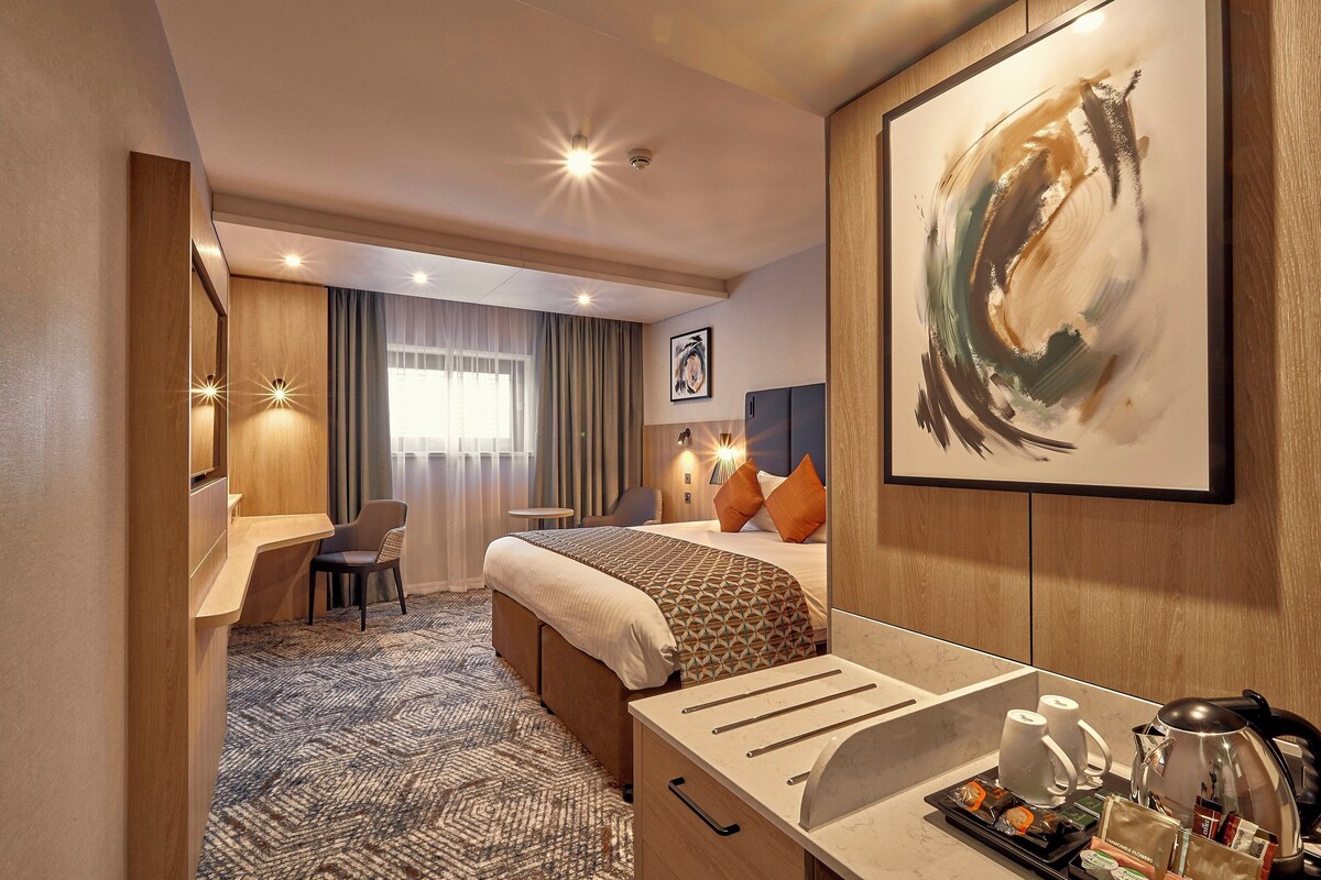 A hotel room with a bed, a desk, a chair, a coffee maker, and a framed abstract painting.