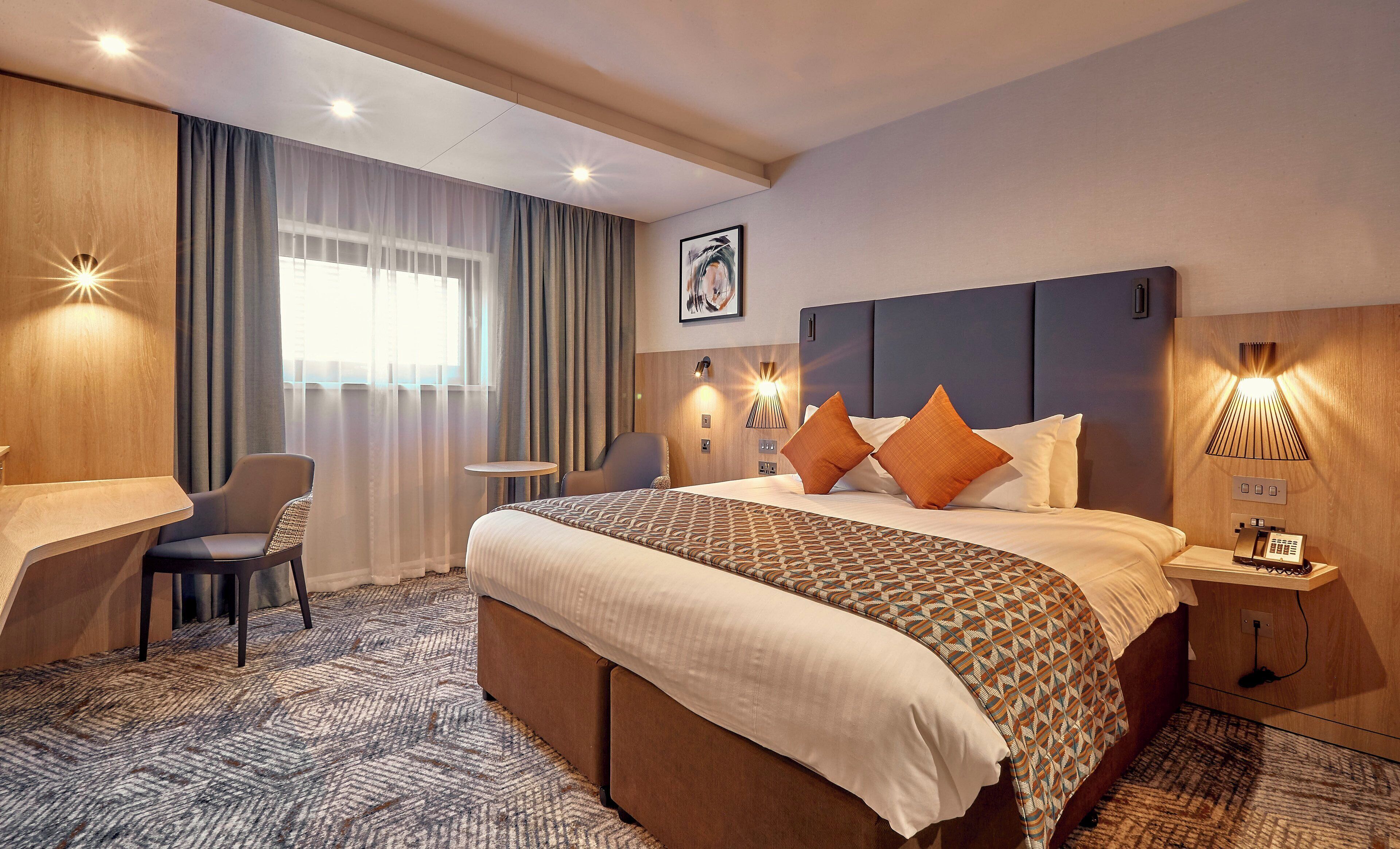 Habitación de hotel con cama, mesita de noche, lámpara, silla, escritorio y ventana con cortinas.