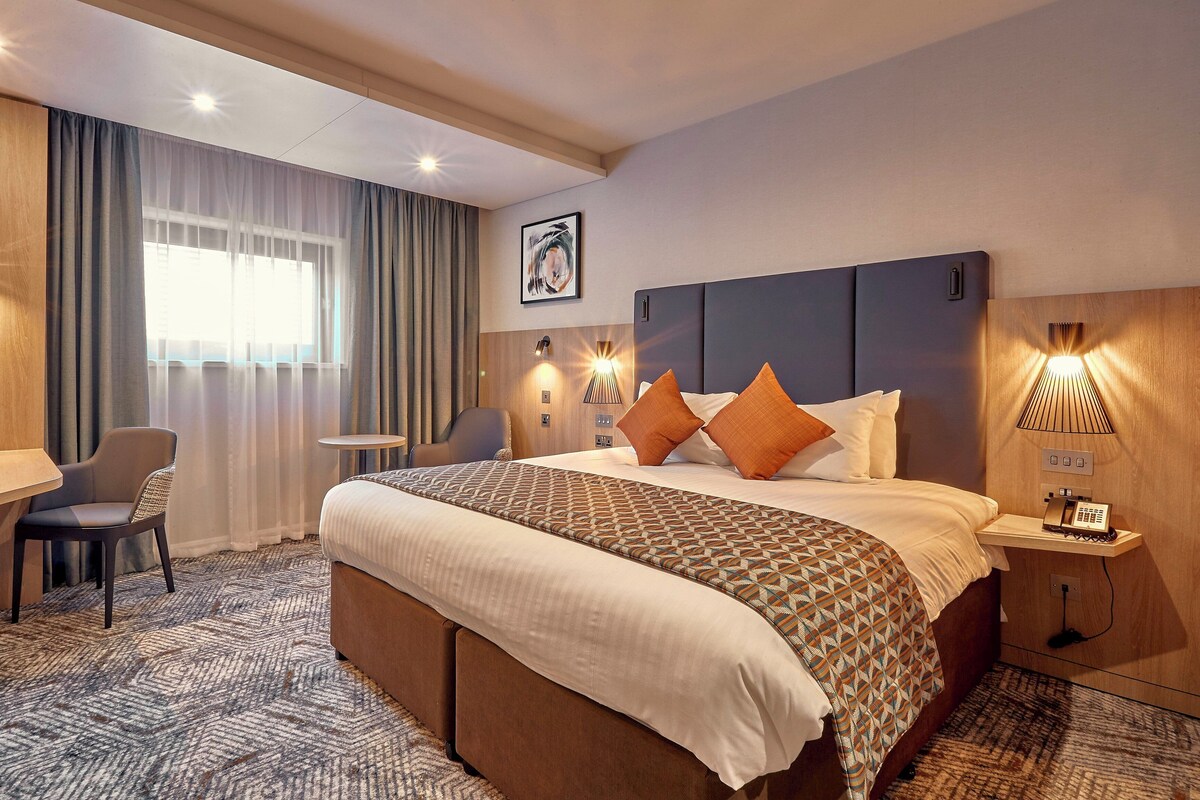 A hotel room with a bed, bedside table, lamp, chair, desk, and a window with curtains.
